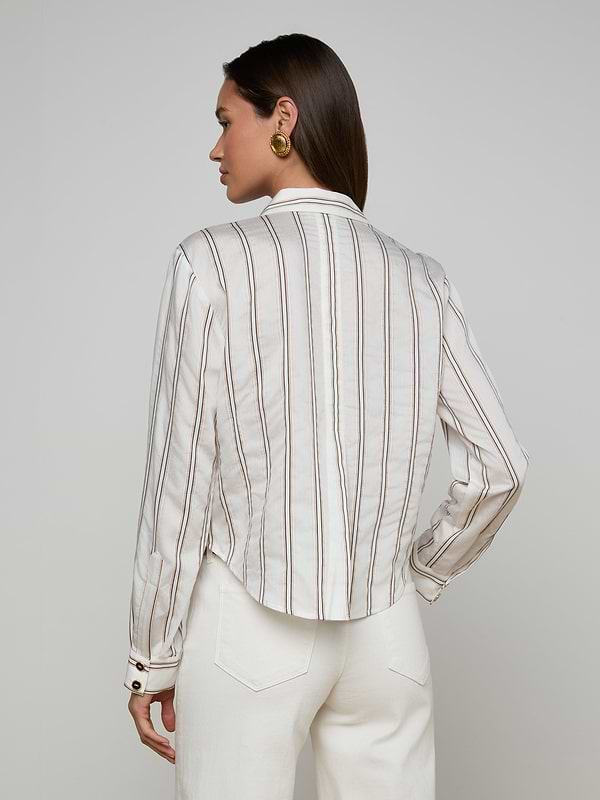 A woman stands facing away, wearing the Cosette Shirt—a tailored white long-sleeve top with vertical jacquard stripes—paired with white pants. She has straight brown hair and accessorizes with gold round earrings, set against a plain, light-colored background.