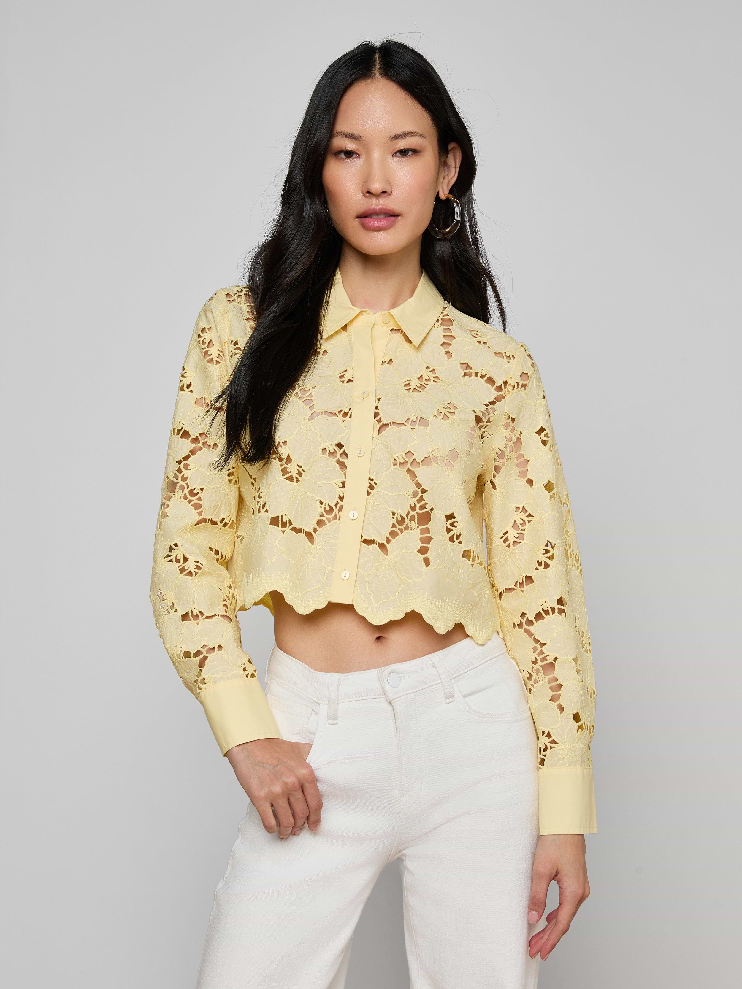 A woman wearing the Seychelle Cropped Eyelet Blouse—a light yellow, long-sleeve poplin lace crop top with a scalloped hem—paired with high-waisted white pants, stands against a plain background.