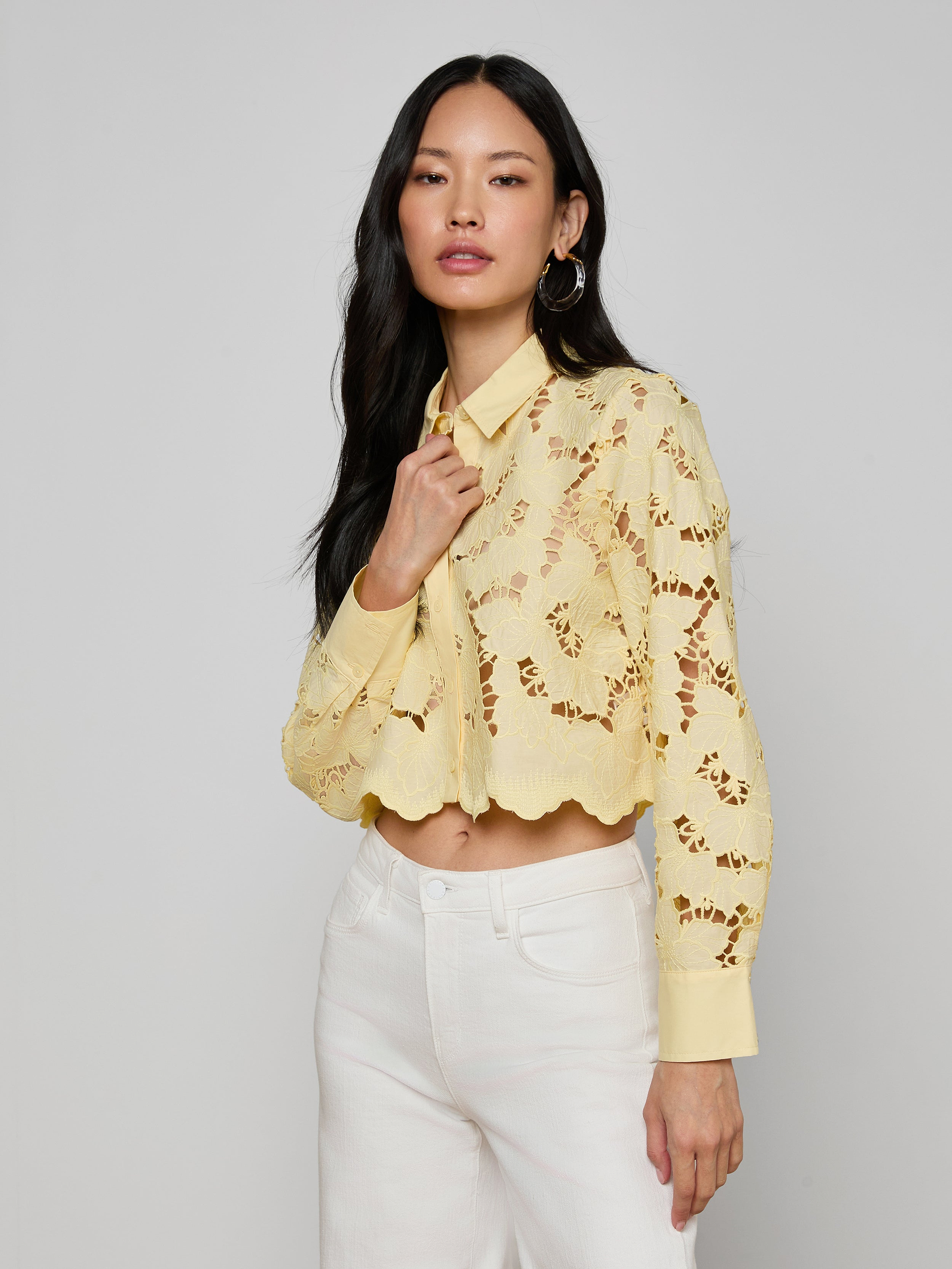 A woman in the Seychelle Cropped Eyelet Blouse—a pale yellow, floral eyelet top with long sleeves—paired with white pants stands against a plain background, looking confidently at the camera.