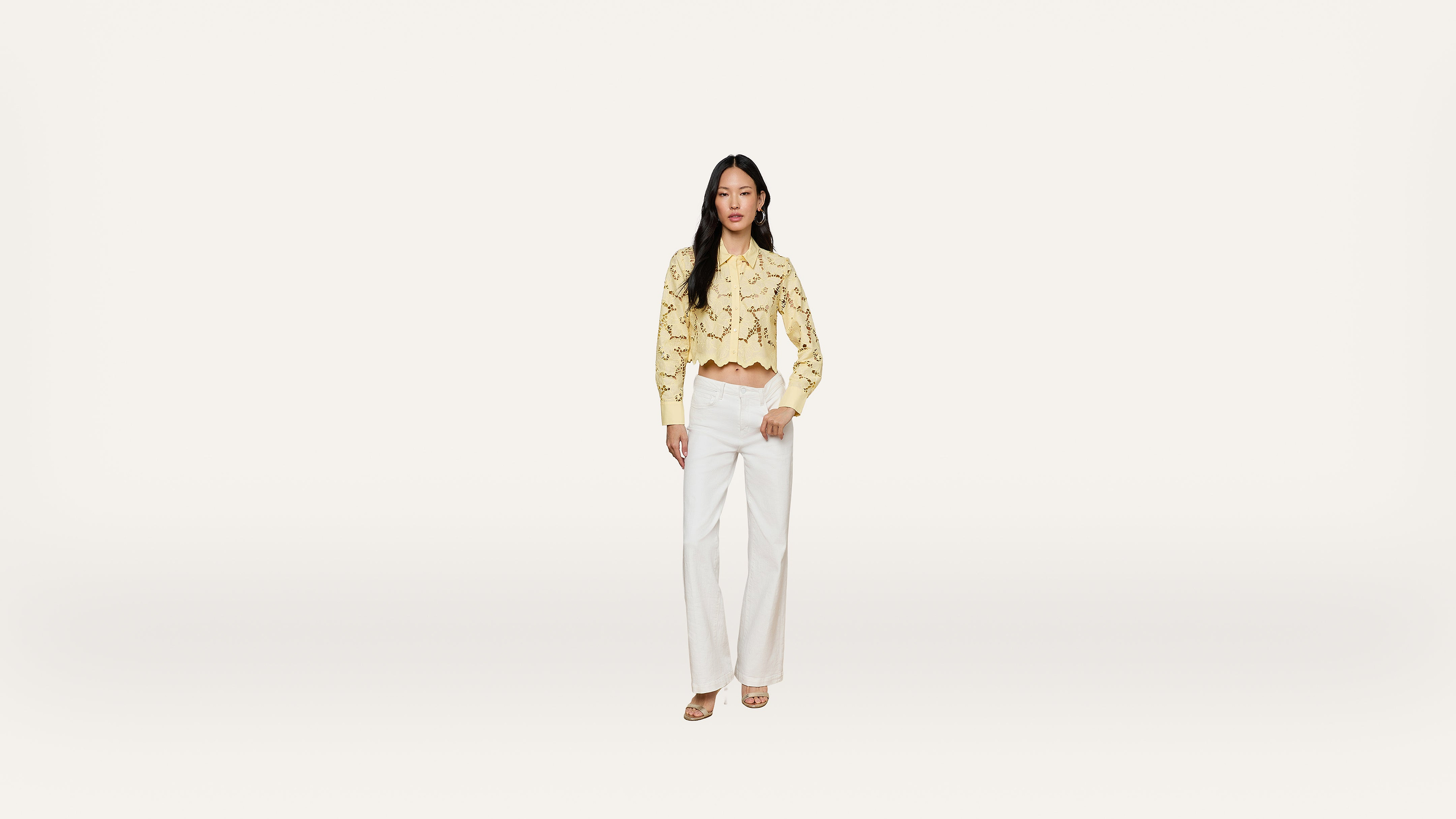 A woman stands against a plain white background, wearing a yellow long-sleeve crop top with lace details and high-waisted white wide-leg pants, completed with light-colored sandals.