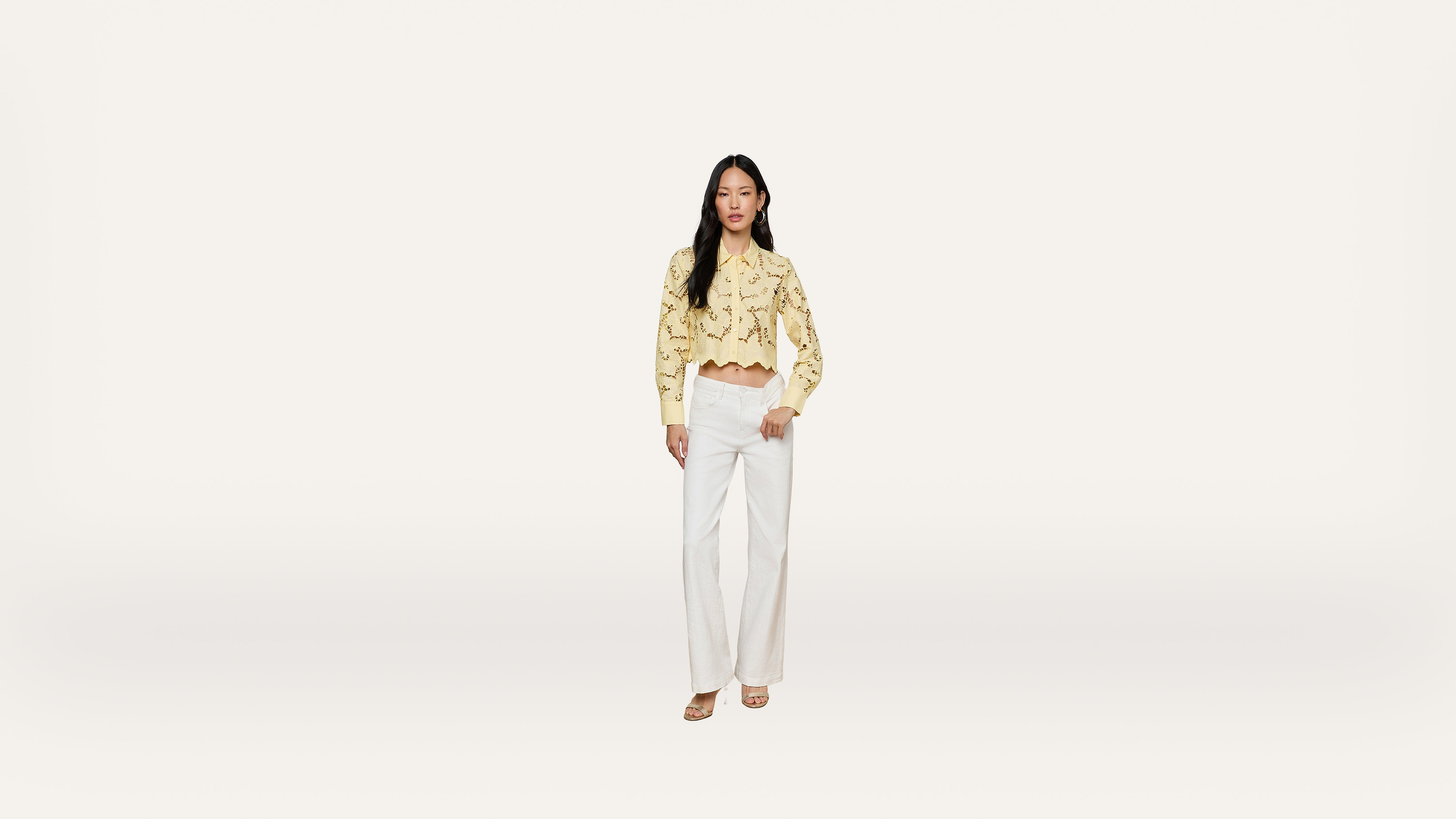 A woman stands against a plain white background, wearing a yellow long-sleeve crop top with lace details and high-waisted white wide-leg pants, completed with light-colored sandals.