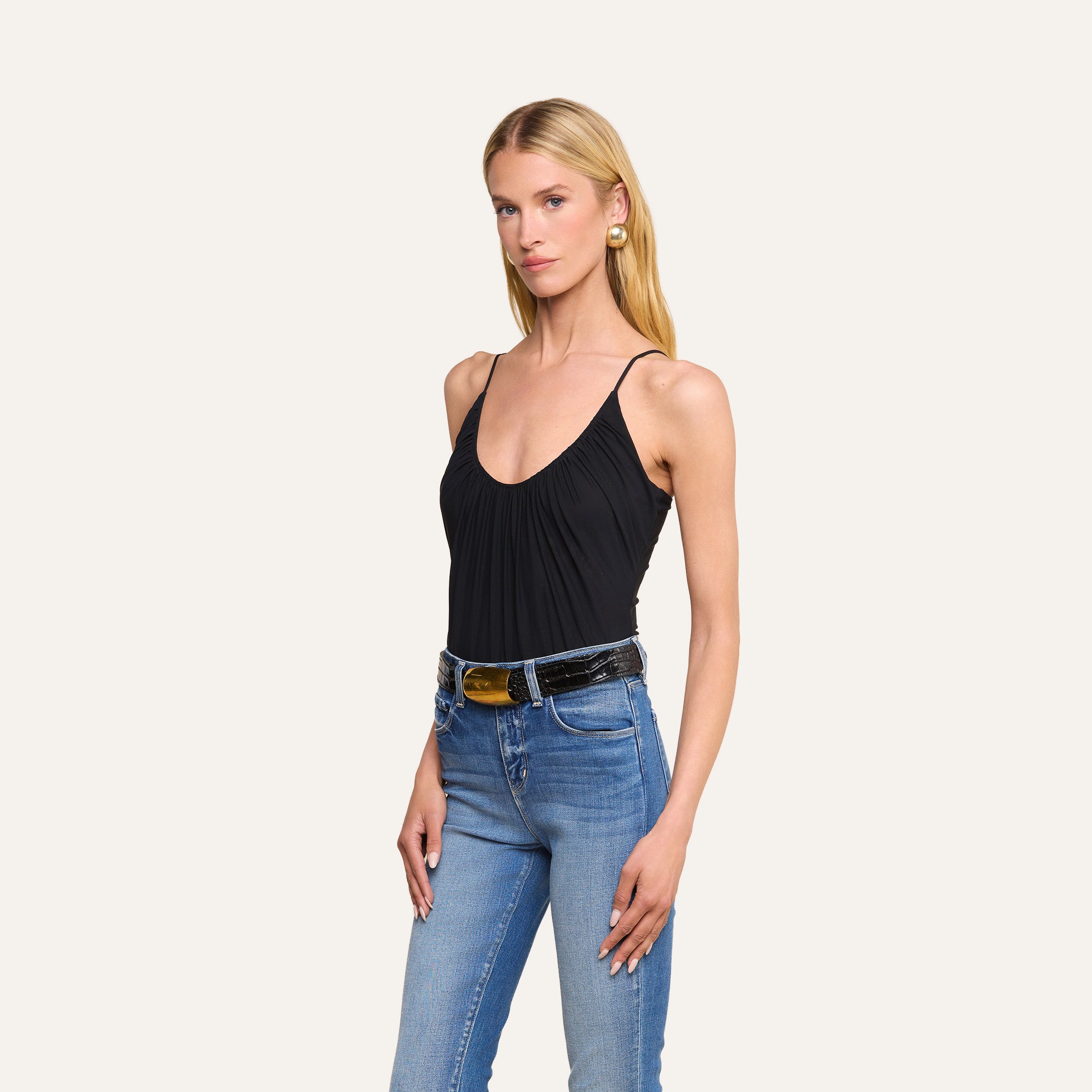 A woman with long blonde hair wears the Gelina Mesh Bodysuit—a black bodysuit with a scoop neckline—paired with blue jeans and a black belt, standing against a plain light background.