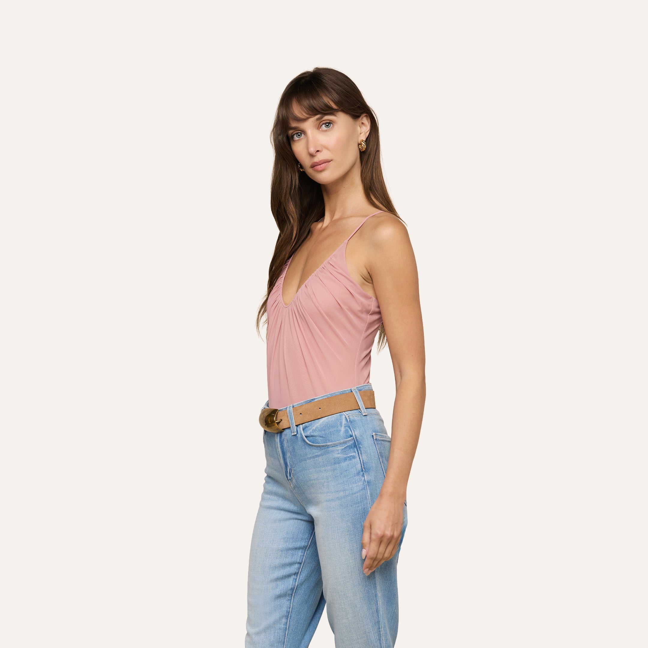 Woman with long brown hair wearing the Gelina Mesh Bodysuit—a pink semi-sheer mesh, sleeveless V-neck design with adjustable straps—paired with light blue jeans and a brown belt, standing against a plain light background and looking slightly to the side.