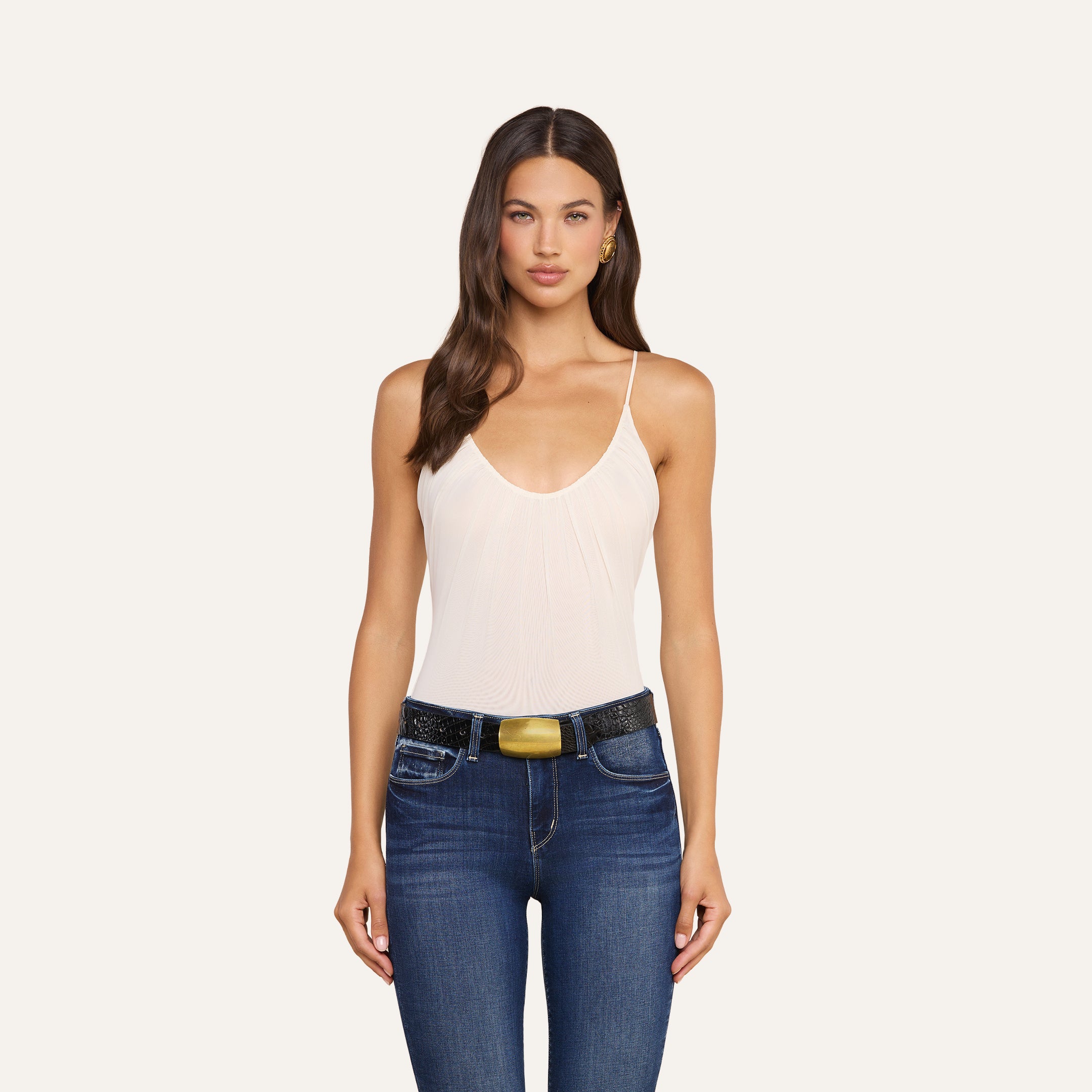 A woman with long brown hair wears the Gelina Mesh Bodysuit—a sleeveless, light pink bodysuit with adjustable straps—paired with blue jeans and a black belt. She stands facing forward against a plain white background.