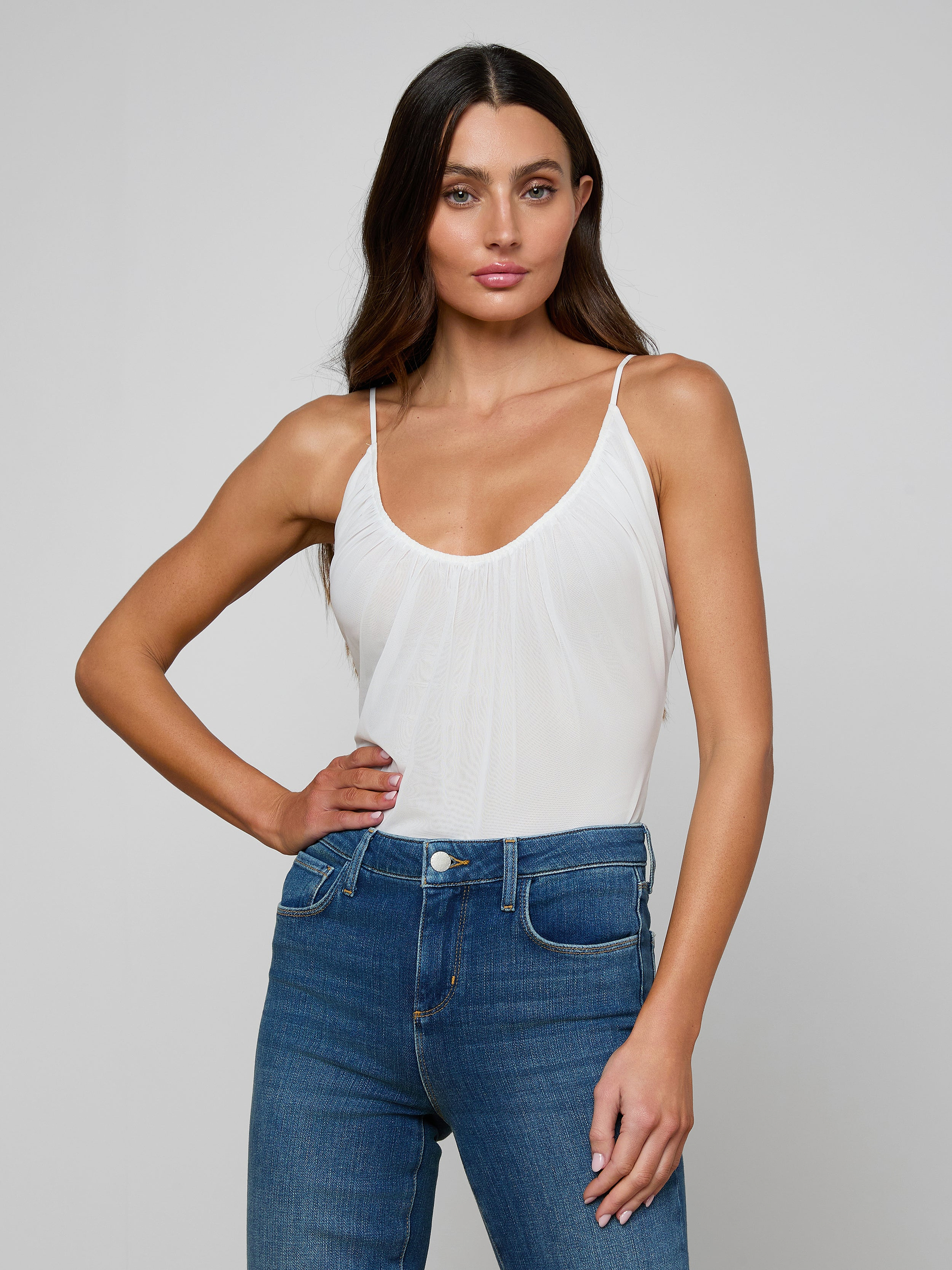 A woman with long brown hair wears the Gelina Ruched Bodysuit, featuring a white mesh design and adjustable straps, paired with blue jeans as she poses with one hand on her hip against a plain light background.