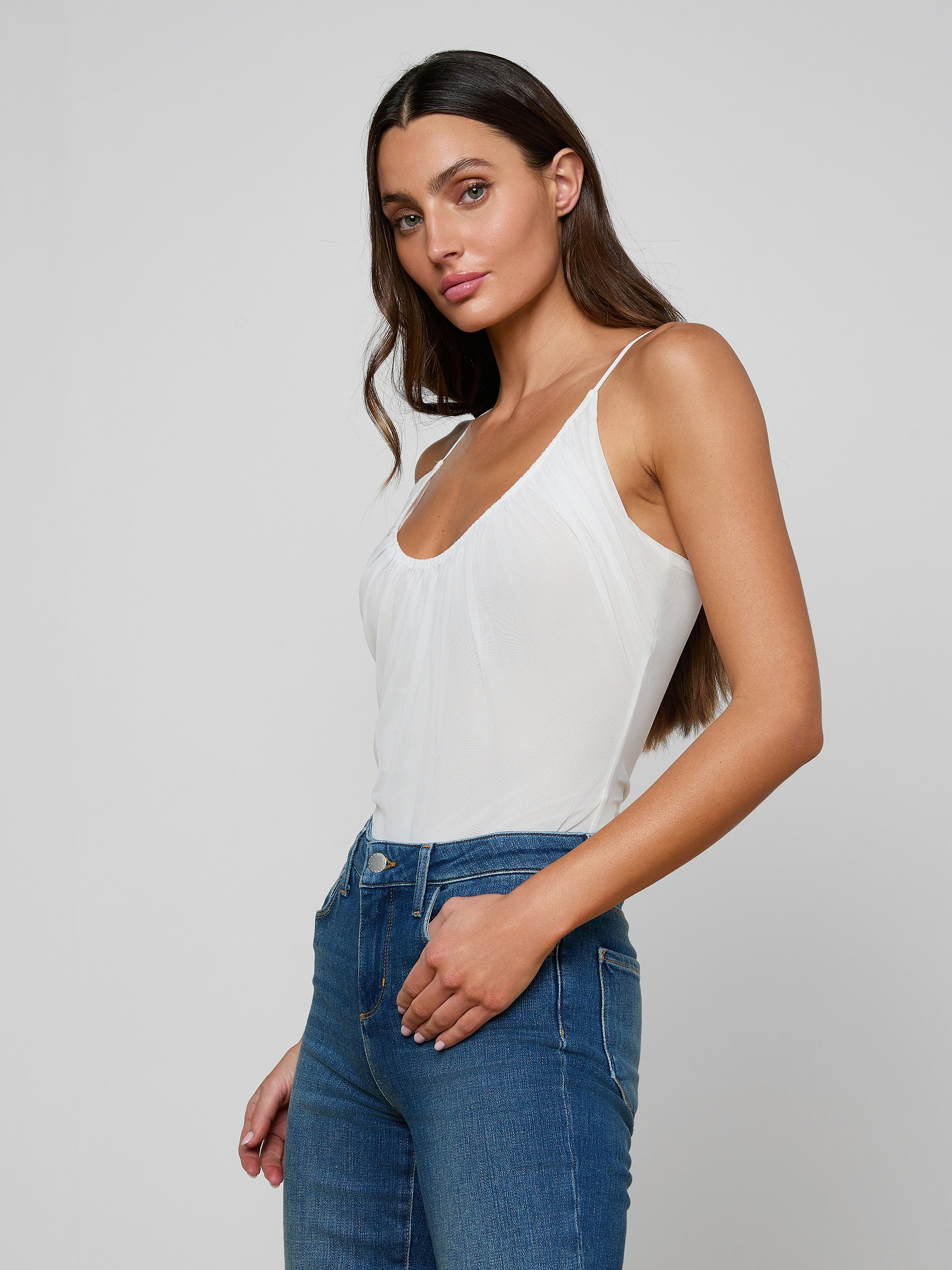 A woman with long dark hair stands confidently against a plain light background, wearing the Gelina Ruched Bodysuit with adjustable straps and blue jeans. She poses in a relaxed manner, looking directly at the camera.