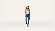 A woman wearing a white tank top, blue cropped jeans, and black high-heeled sandals stands against a plain white background, looking slightly to her left.