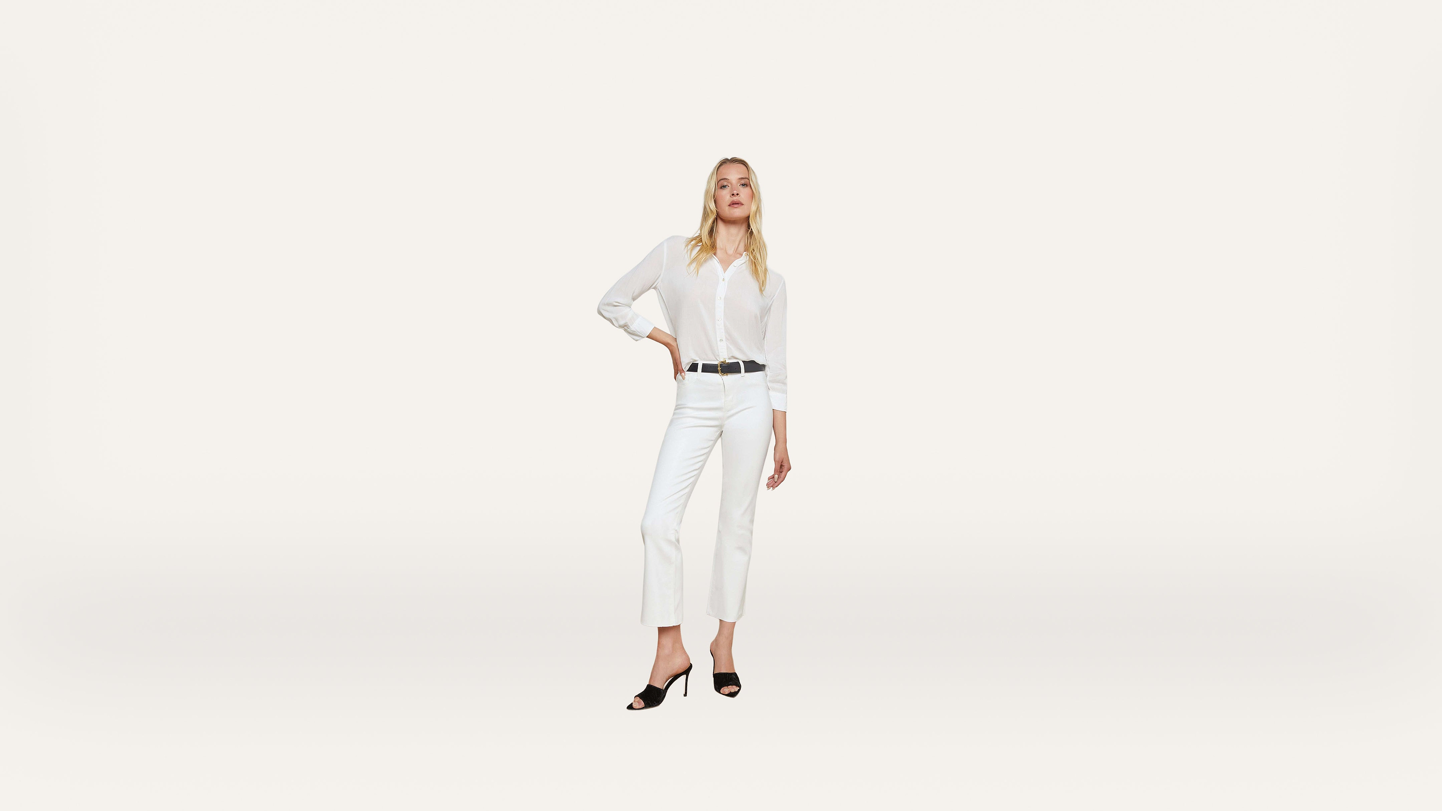 Woman standing confidently in all-white attire, including a button-up shirt and cropped pants, with black heels and a black belt, against a plain white background.