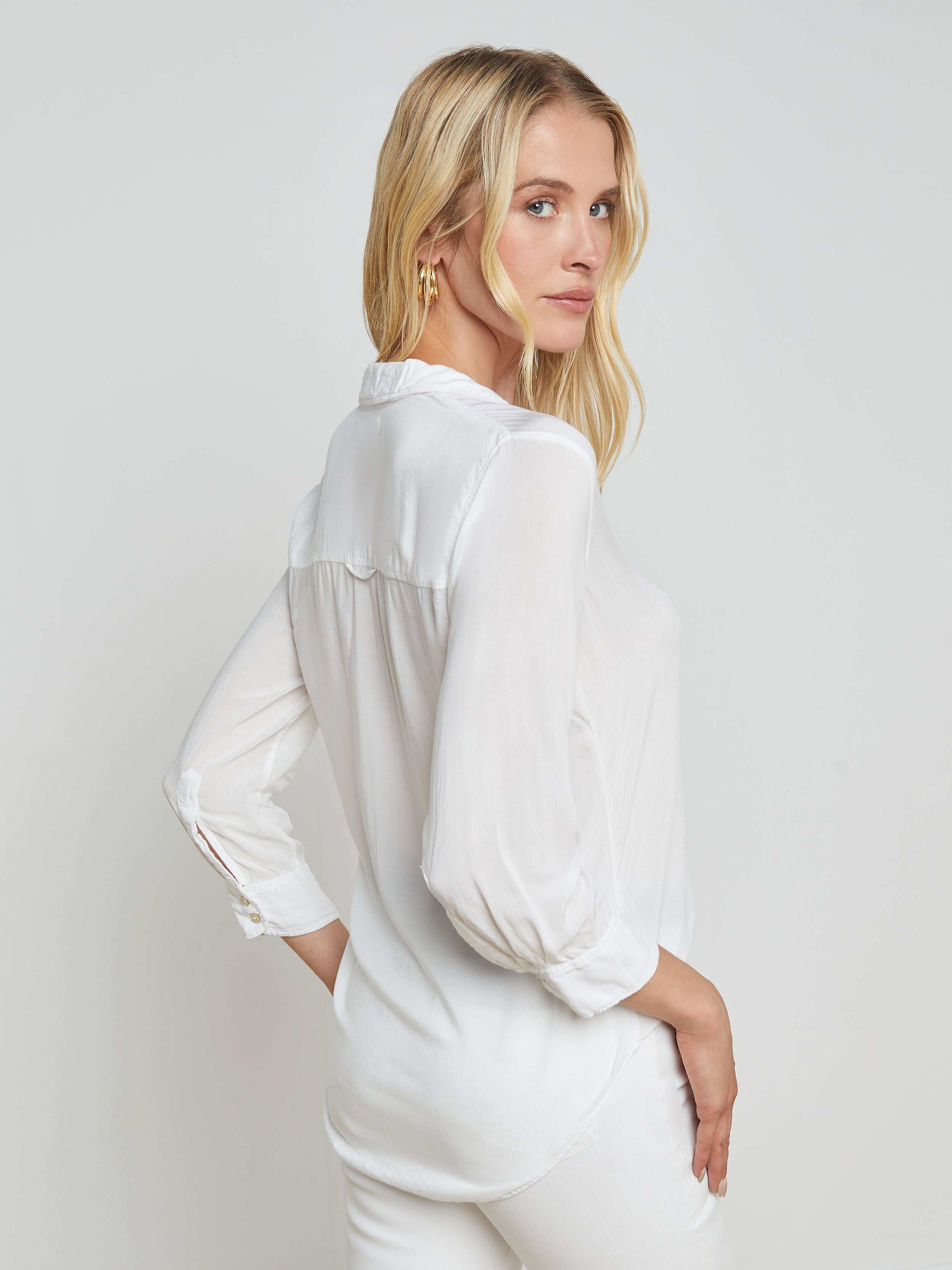 A woman with long blonde hair wears the Marlene Blouse and white pants, standing sideways and looking over her shoulder against a plain light background.