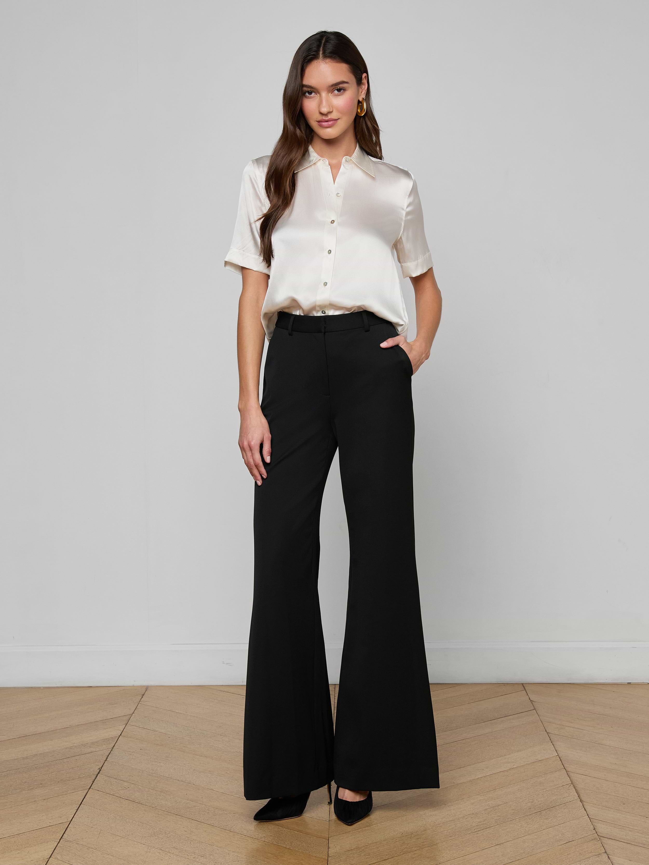 A woman stands against a plain white background, wearing the Ellah Silk Blouse with short sleeves, cream pants, a belt, and brown high heels. She has long brown hair and a neutral expression.