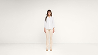 A woman with long brown hair stands in a minimalist, white room. She wears a white button-up shirt, beige pants, and tan shoes, facing forward with a neutral expression.