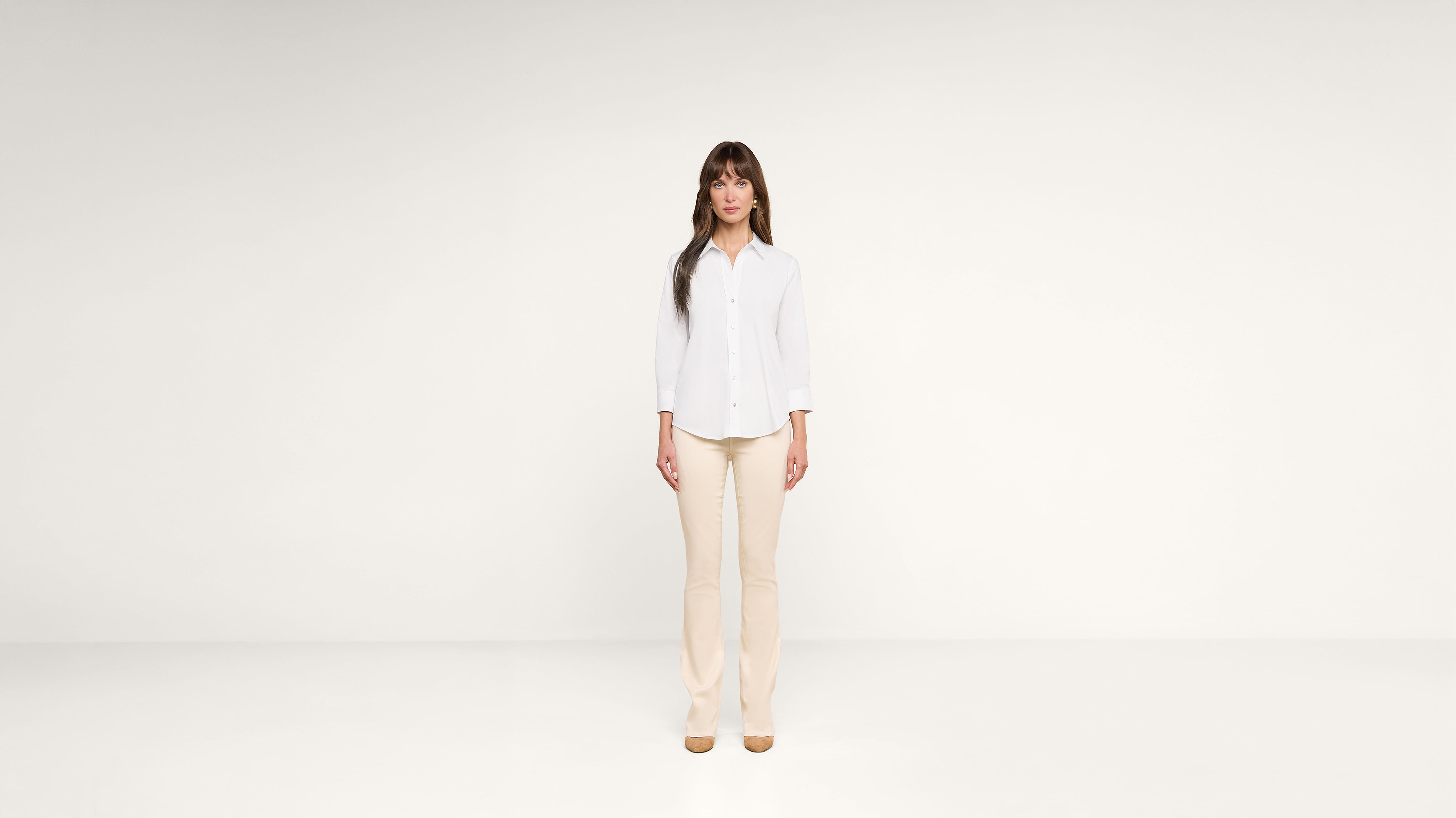 A woman with long brown hair stands in a minimalist, white room. She wears a white button-up shirt, beige pants, and tan shoes, facing forward with a neutral expression.