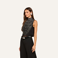 A woman stands against a plain light background wearing the Yun Draped Crystal Tank—an embellished tank adorned with layered silver crystals—paired with black pants, a black belt with a silver buckle, and silver earrings.
