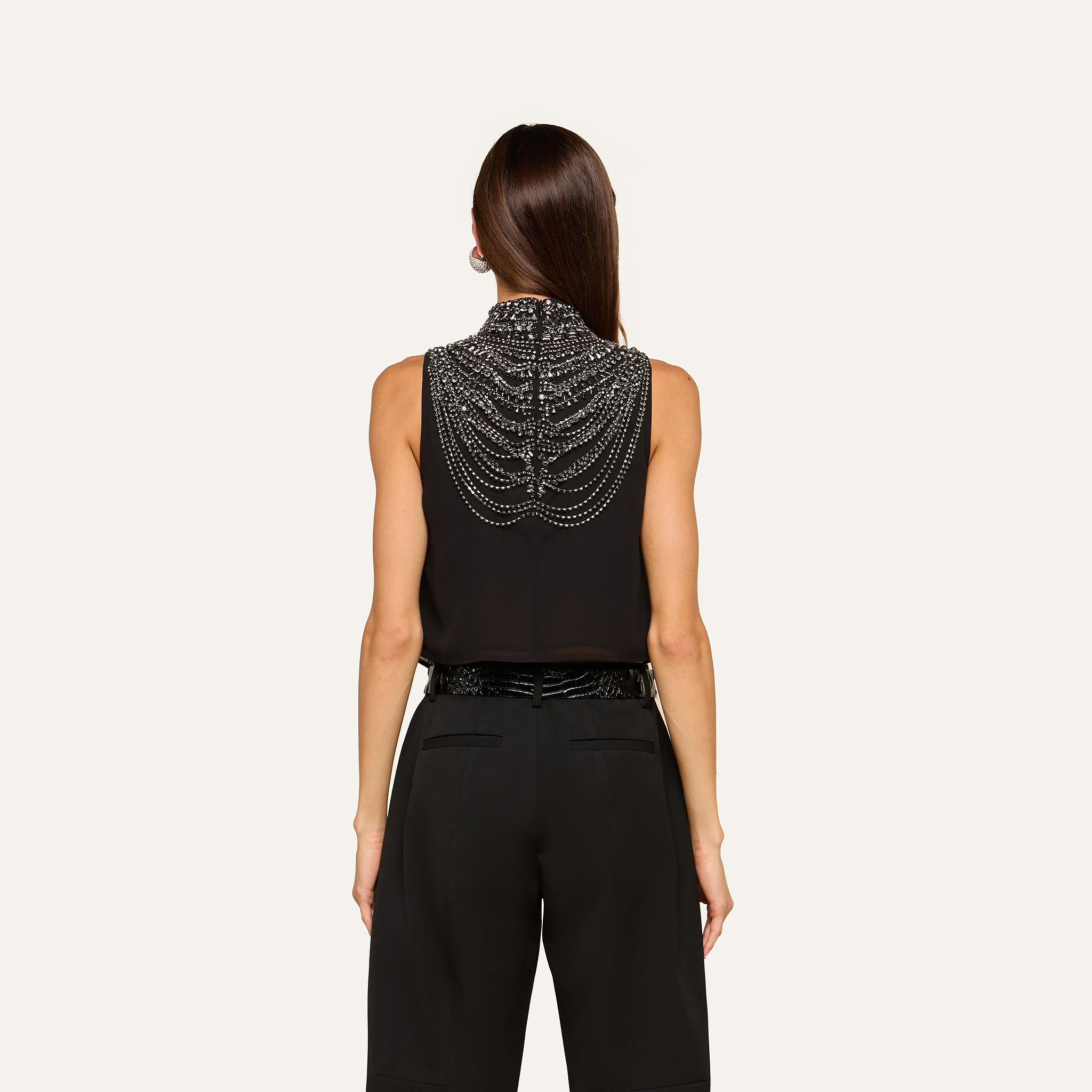 A person with straight brown hair wears the Yun Draped Crystal Tank—a sleeveless black blouse adorned with intricate beaded chains draping across the back—styled as an elegant evening top and paired with high-waisted black pants, shown from behind against a plain light background.
