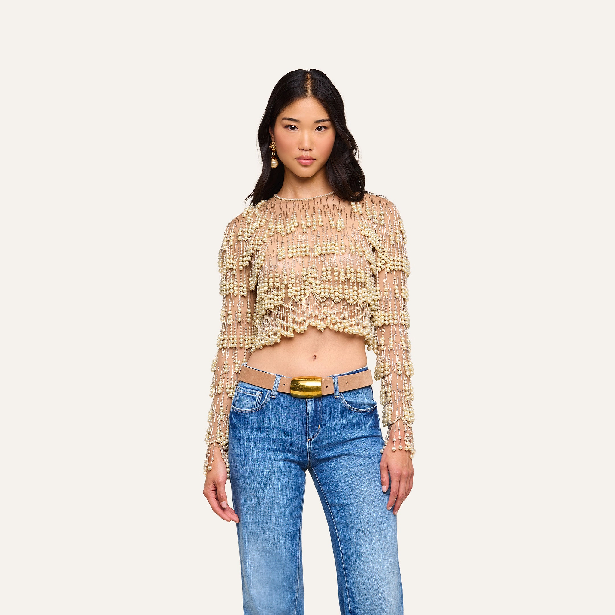 A woman in the Rogan Pearl Fringe Top—a beige, mesh, long sleeve cropped party shirt with pearl fringe—paired with blue jeans stands against a plain, light background, facing forward with a neutral expression.