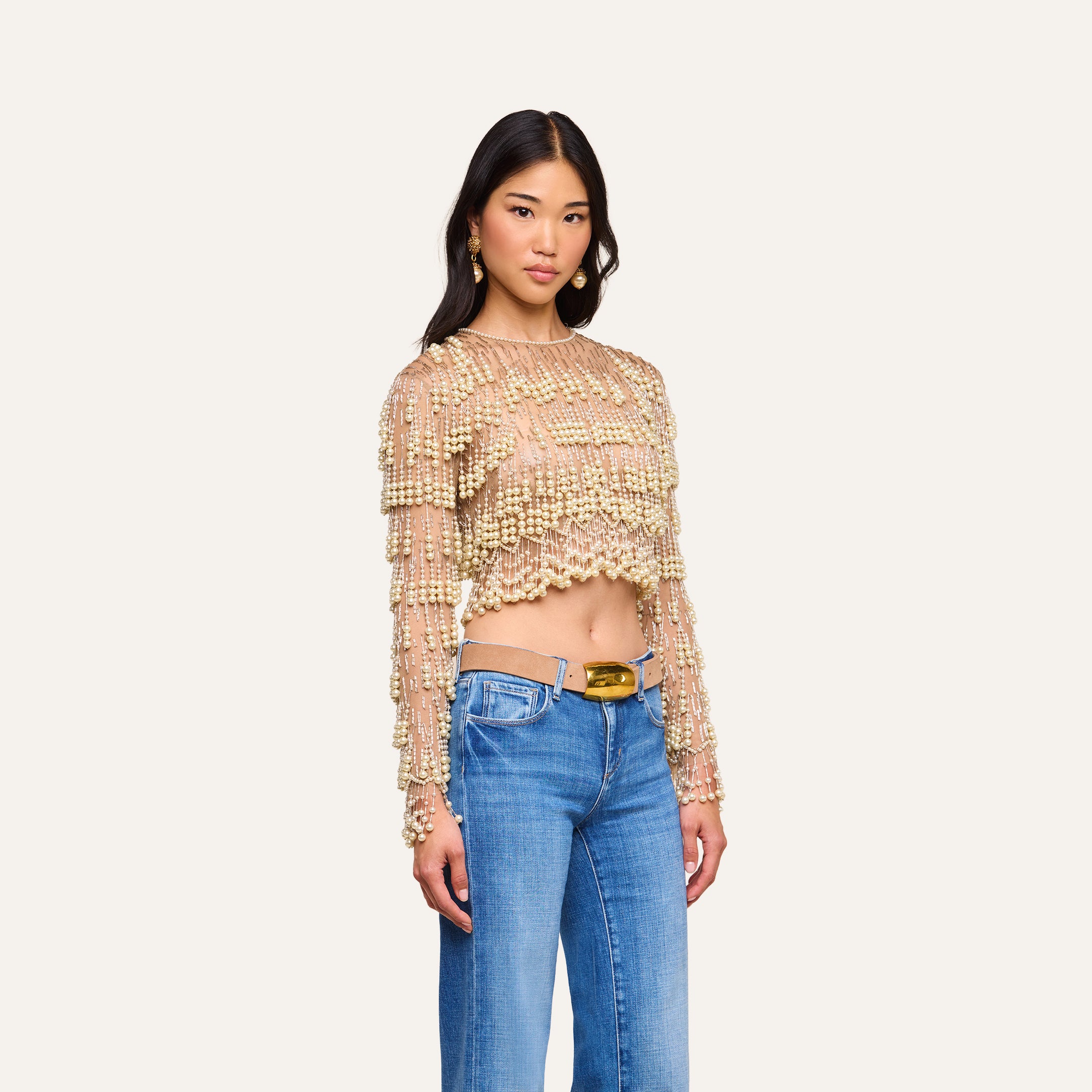 Wearing the Rogan Pearl Fringe Top—a sheer mesh, long sleeve cropped party top embellished with gold beads—paired with blue jeans and gold earrings, a woman stands against a plain light background.