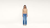 A woman stands facing forward against a plain white background, wearing a long-sleeve, multicolored crop top, high-waisted blue jeans, a tan belt, and beige shoes.