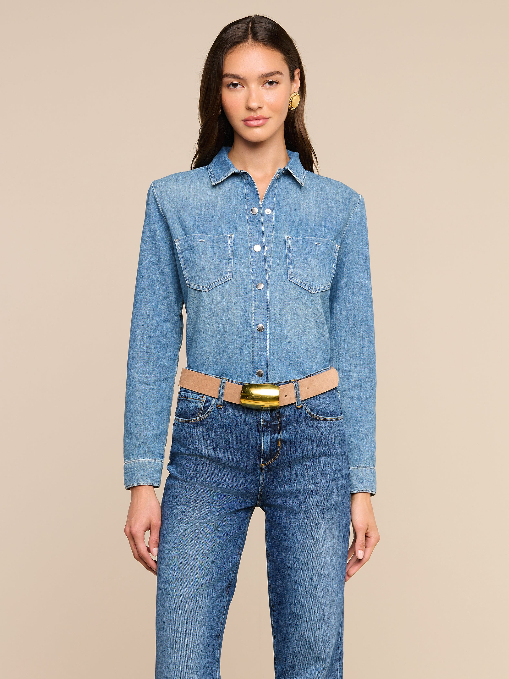 Mujer con el pelo largo y castaño que lleva la camisa «Isla» —una camisa vaquera de botones muy ligera— combinada con unos vaqueros azules de lavado medio y un cinturón beige con hebilla dorada; está de pie frente a un fondo liso beige y mira a la cámara con una expresión neutra.