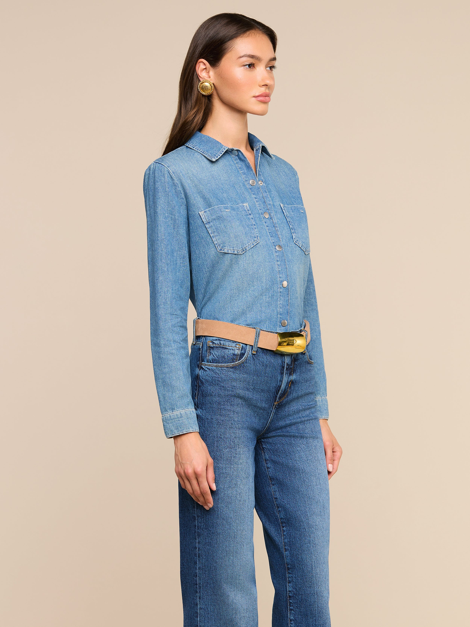 Una mujer que lleva la camisa «Isla» en chambray de tono medio, metida por dentro de unos vaqueros azules de cintura alta y complementada con un cinturón ancho de color beige y unos pendientes dorados, posa frente a un fondo liso de color beige.