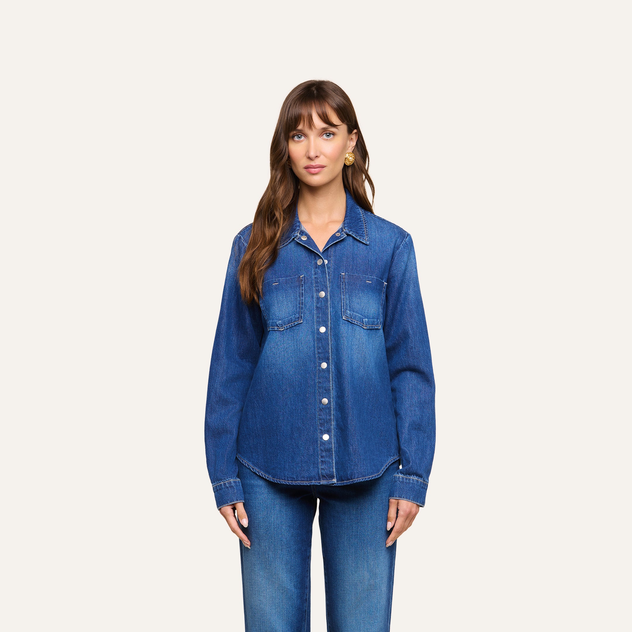 A woman with long brown hair stands facing forward, wearing the Isla Shirt—a featherweight denim shirt with front pockets—and matching blue jeans, against a plain light background.