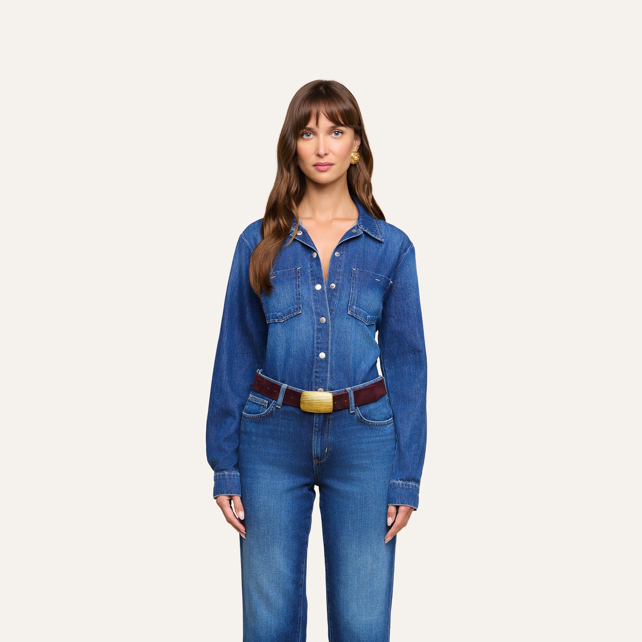 A woman with long brown hair is wearing the Isla Shirt, a lightweight denim top, paired with matching jeans and accessorized with a brown belt featuring a large light-colored buckle, standing against a plain light background.