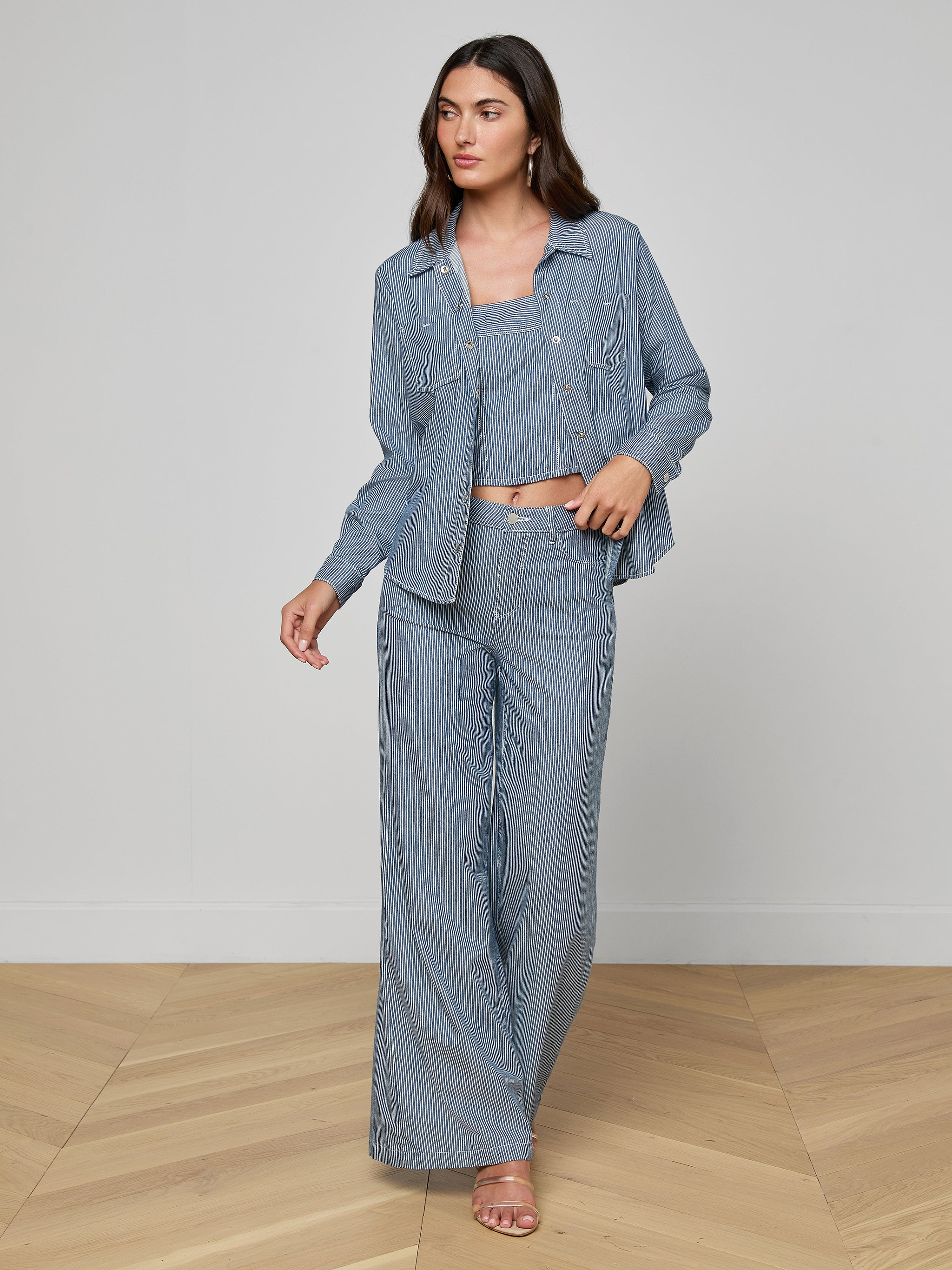 A woman stands against a plain, light background wearing the Isla Shirt in featherweight light blue denim, paired with a matching crop top and wide-leg pants. She looks off to the side.