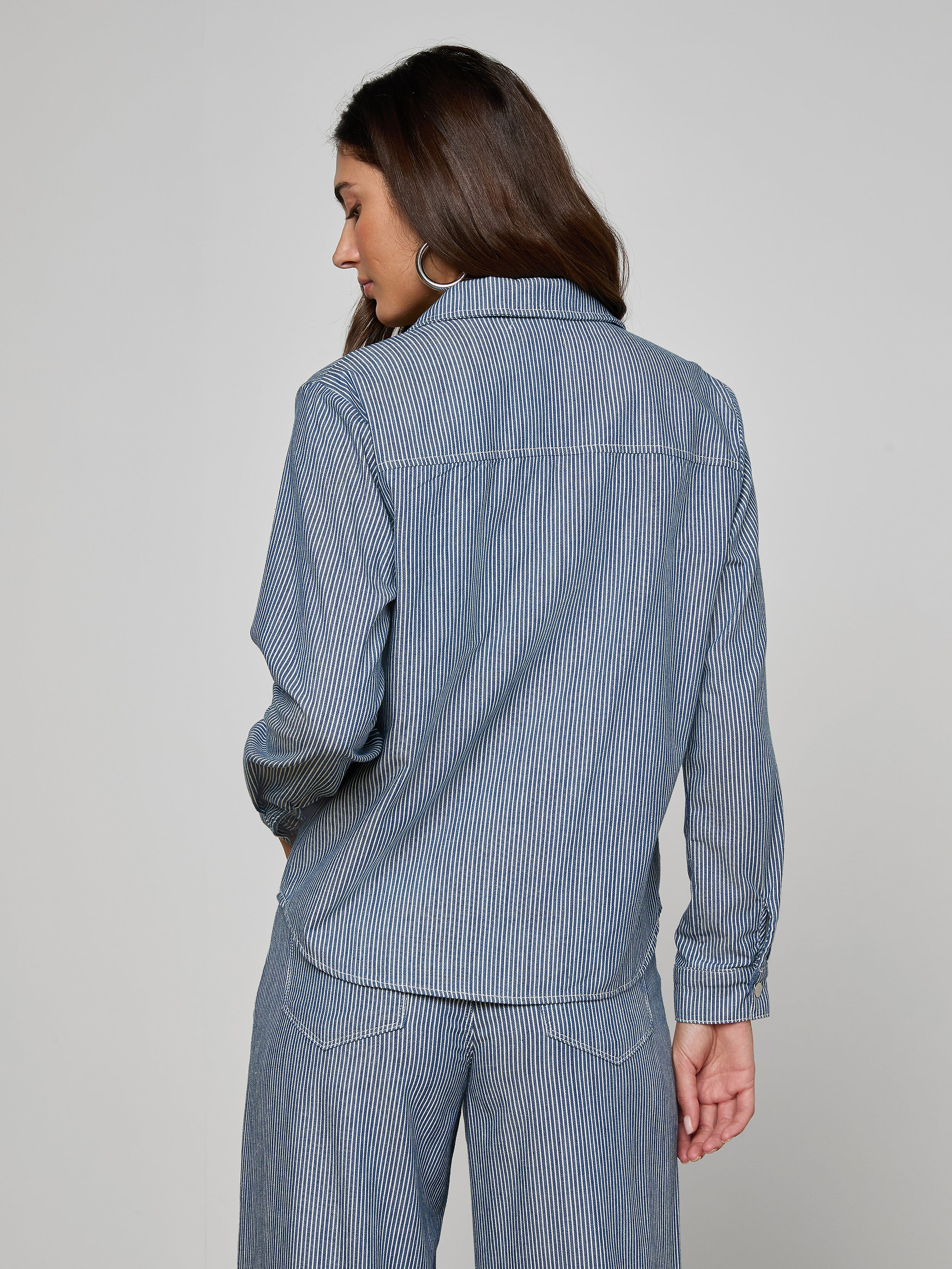 Woman standing with her back to the camera, wearing the Isla Shirt in indigo pinstripe featherweight denim from L’AGENCE, paired with matching pants, against a plain light background.