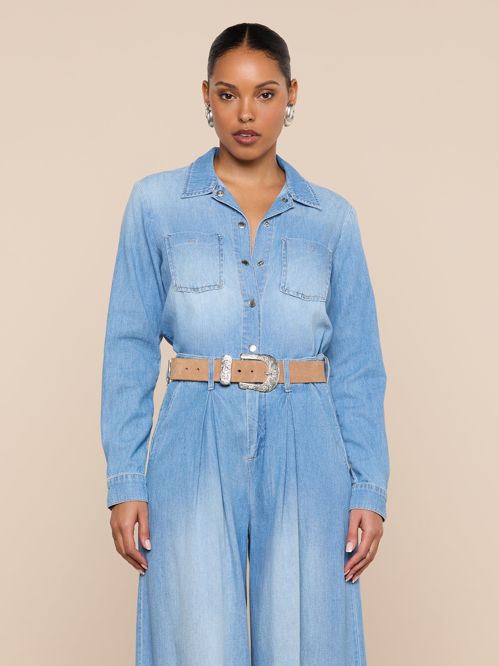 A woman stands facing forward against a plain off-white background, wearing the Isla Shirt styled as a lightweight, vintage-wash denim jumpsuit with a beige belt, hoop earrings, and her hair pulled back.