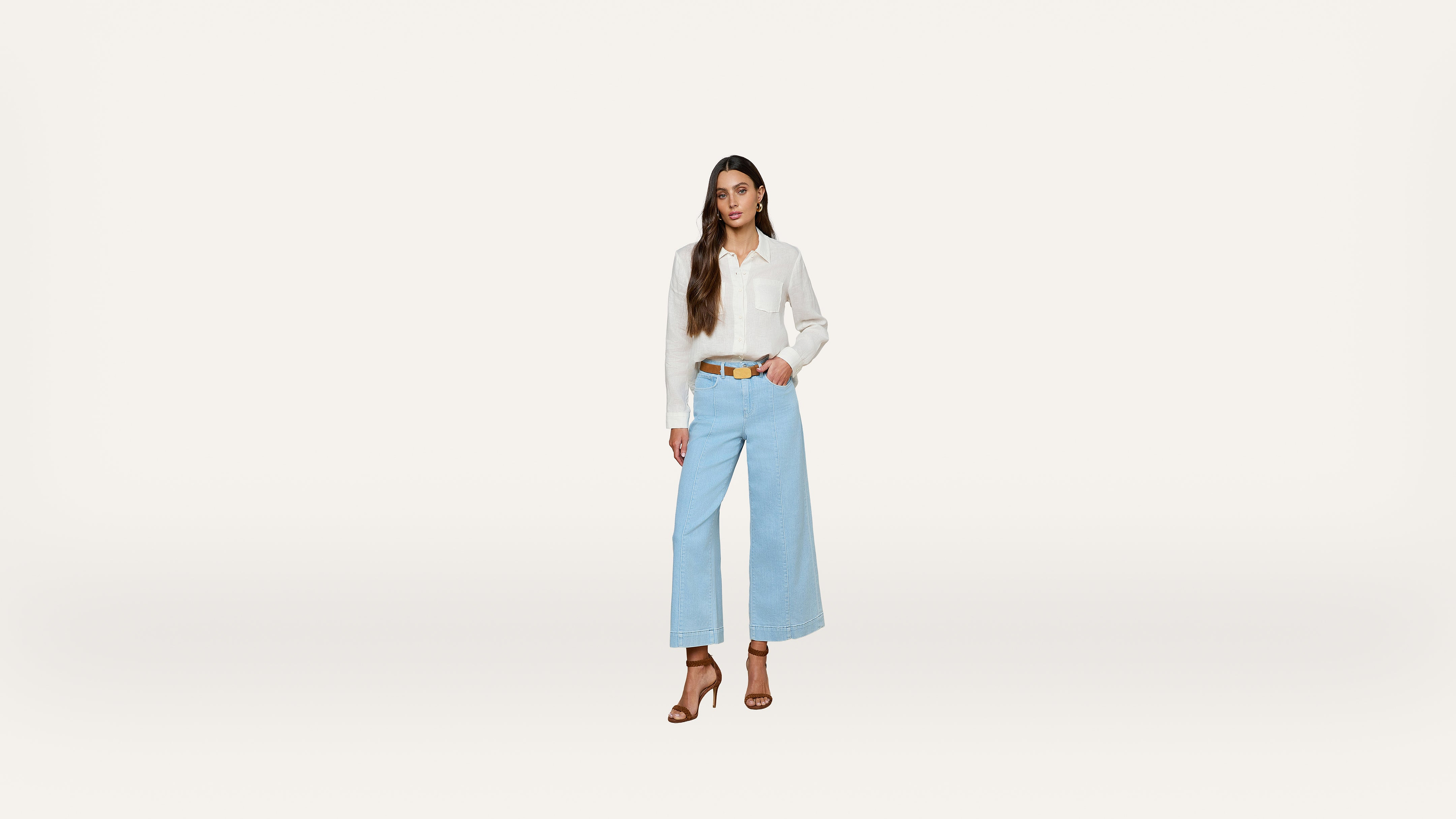 A woman stands against a plain white background, wearing a white blouse, light blue wide-leg jeans, brown belt, and brown heeled sandals, with long brown hair styled in loose waves.