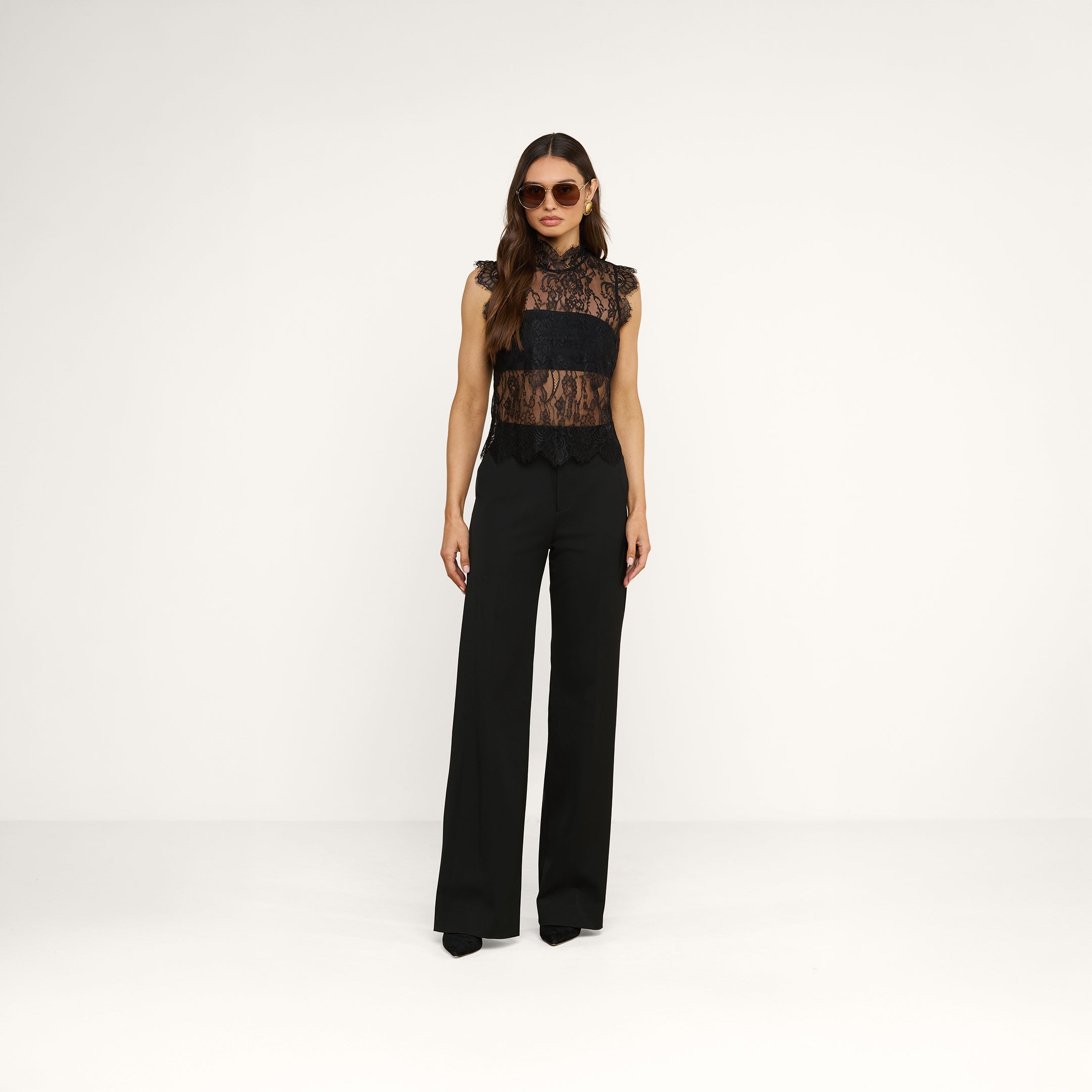 A woman wearing sunglasses, the Tours Lace Top, and high-waisted black pants stands against a plain white background.