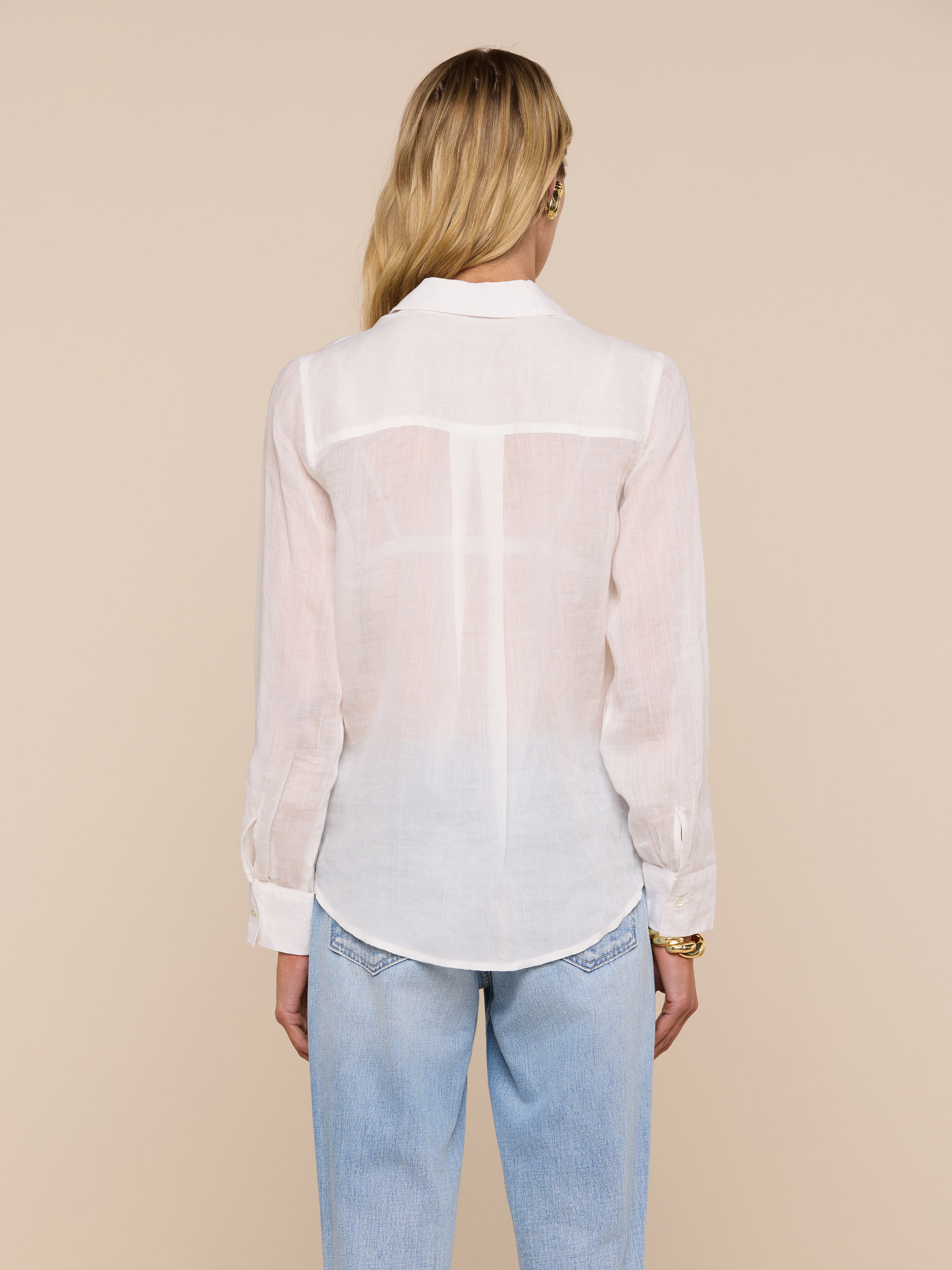 A person with long blonde hair, wearing the Ellery Blouse and light blue jeans, is standing with their back to the camera against a plain light background.