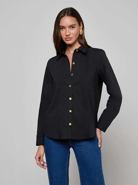 A woman wearing the Ellery Button-Down Shirt, a black stretch cotton blouse with gold buttons, paired with blue jeans stands against a plain light background, looking at the camera with one hand on her hip.