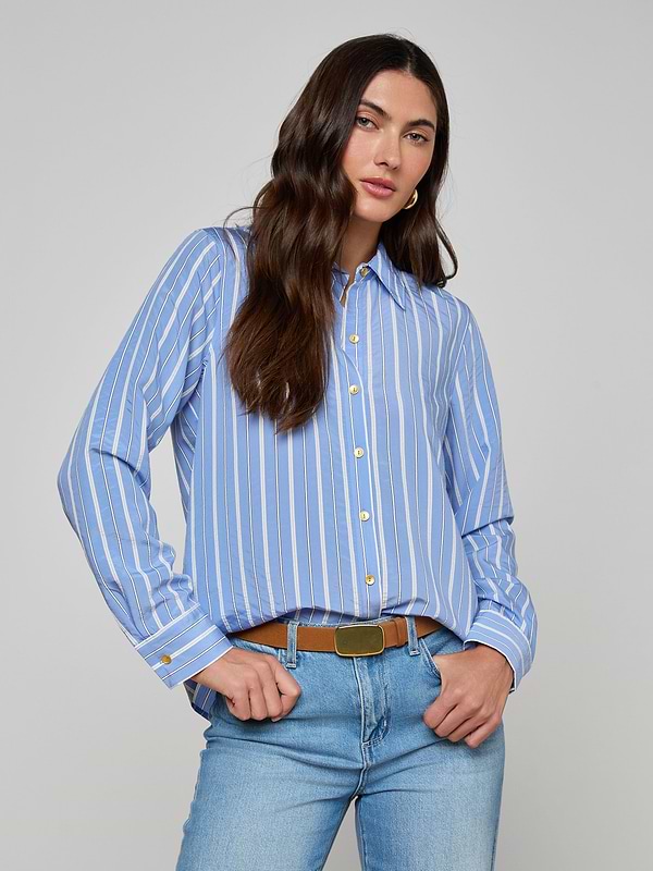 A woman with long dark hair stands against a plain background, hands in her pockets, wearing the Ellery Poplin Shirt— a blue and white striped button-down, tucked into light blue jeans.