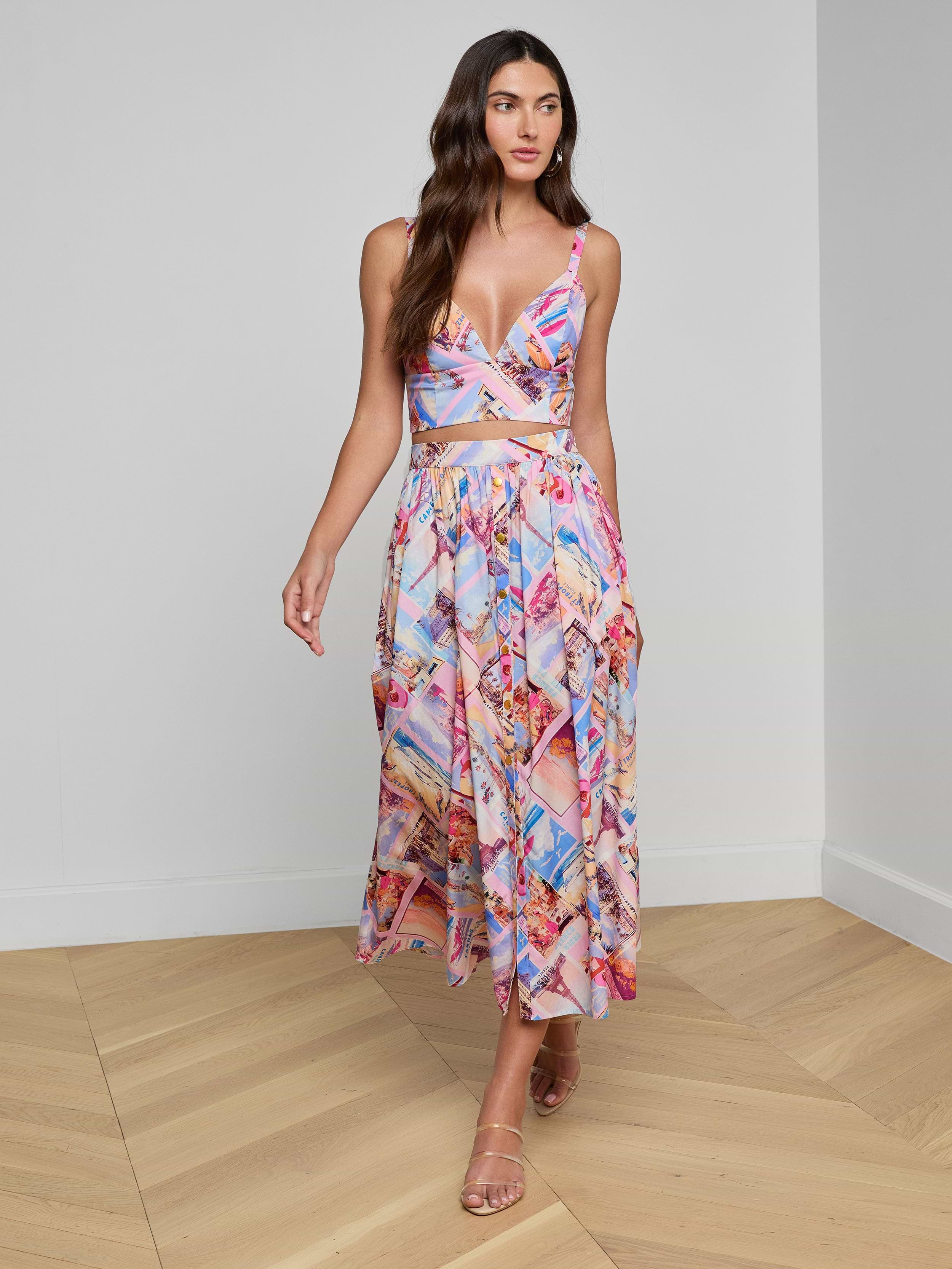 A woman styled in the Kaori Bralette Top and a vibrant postcard print midi skirt, both crafted from cotton-blend poplin, paired with nude heels as she walks against a plain white background.