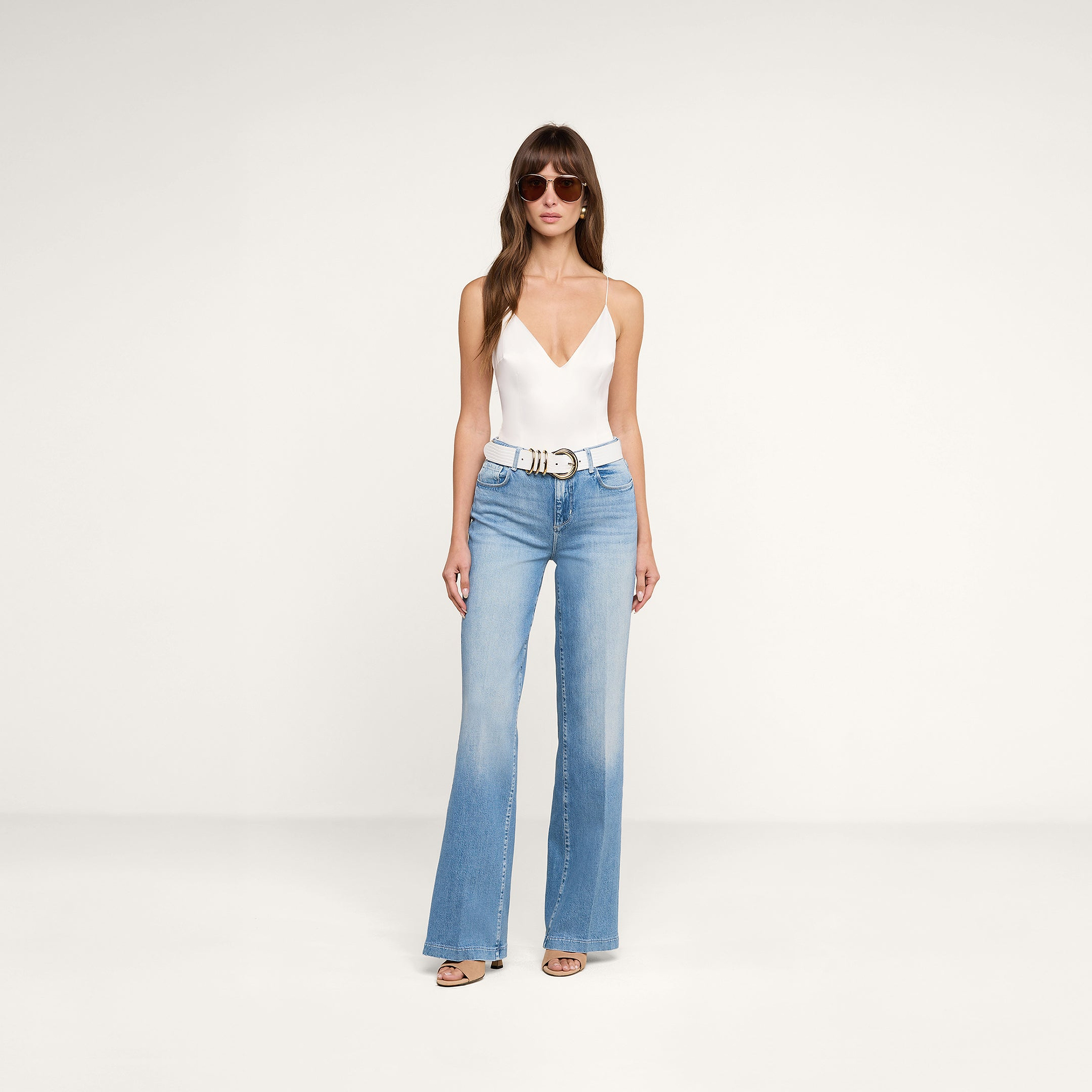 A woman wearing sunglasses, the Dara Stretch Satin Bodysuit—a white sleeveless V-neck top with adjustable straps and a satin base layer—paired with light blue high-waisted flared jeans, a white belt, and sandals, stands against a plain white background.