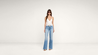 Woman wearing sunglasses, a white deep V-neck sleeveless top, light blue high-waisted wide-leg jeans, and sandals stands against a plain white background.