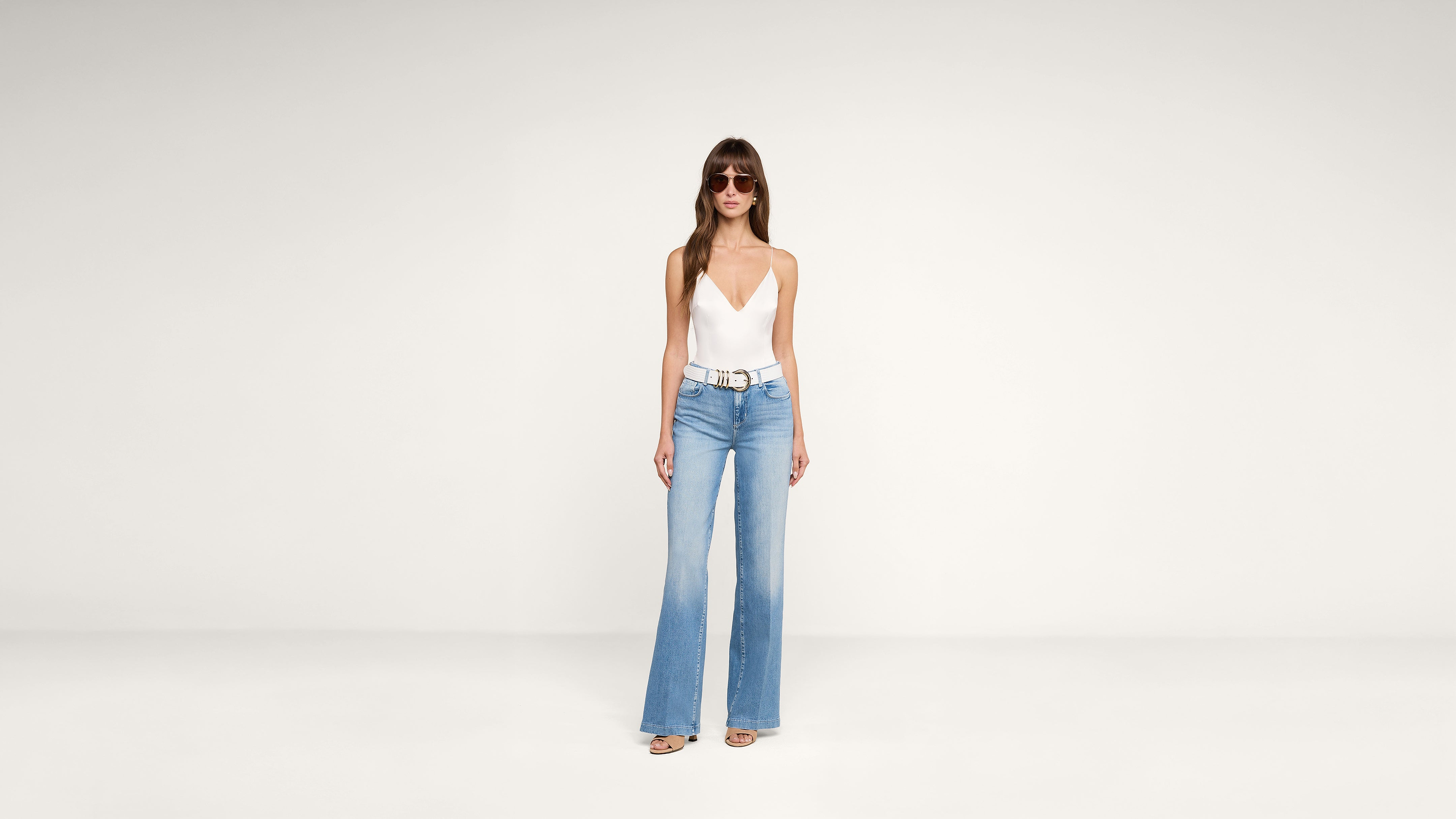 Woman wearing sunglasses, a white deep V-neck sleeveless top, light blue high-waisted wide-leg jeans, and sandals stands against a plain white background.
