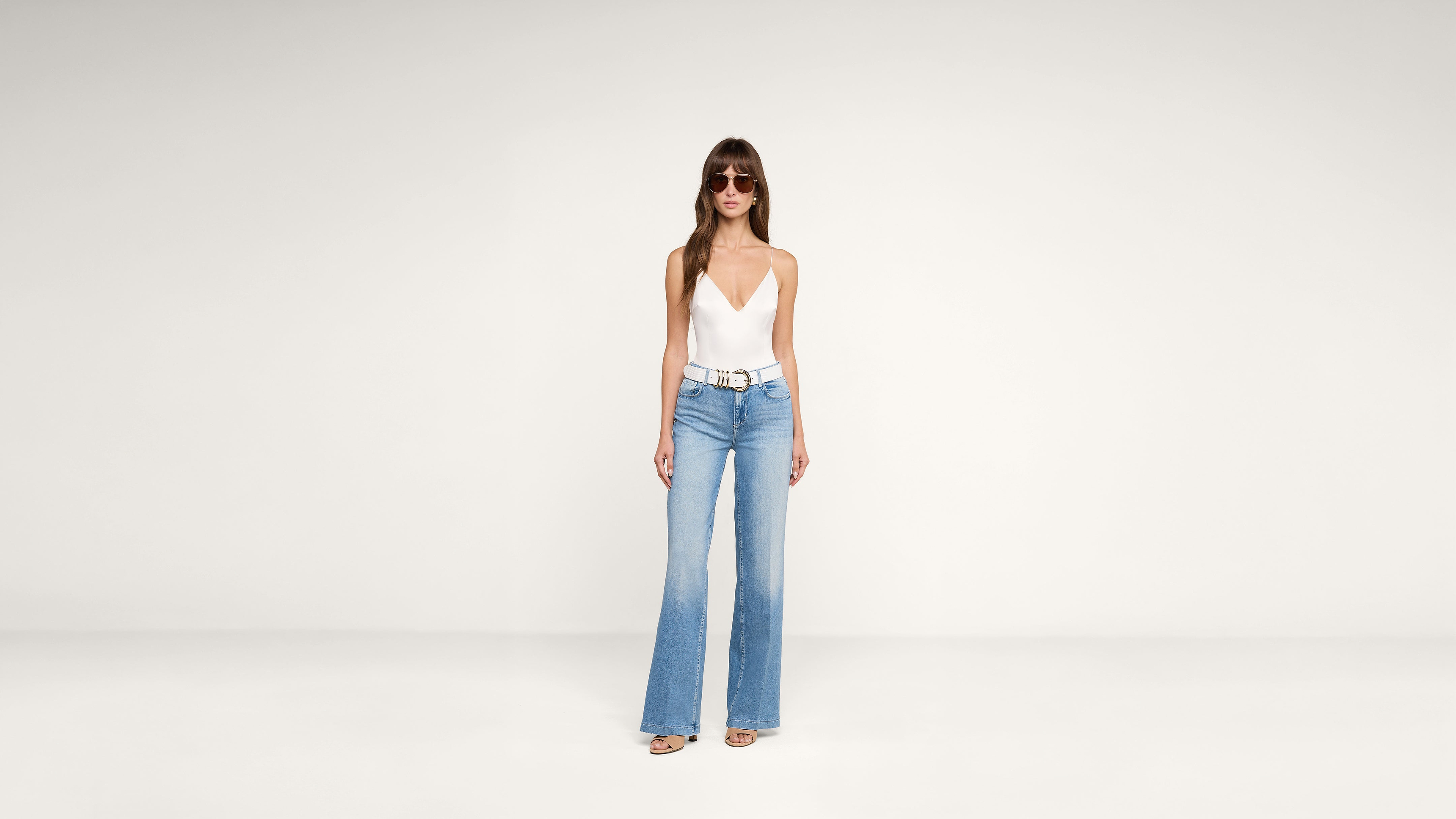 Woman wearing sunglasses, a white deep V-neck sleeveless top, light blue high-waisted wide-leg jeans, and sandals stands against a plain white background.