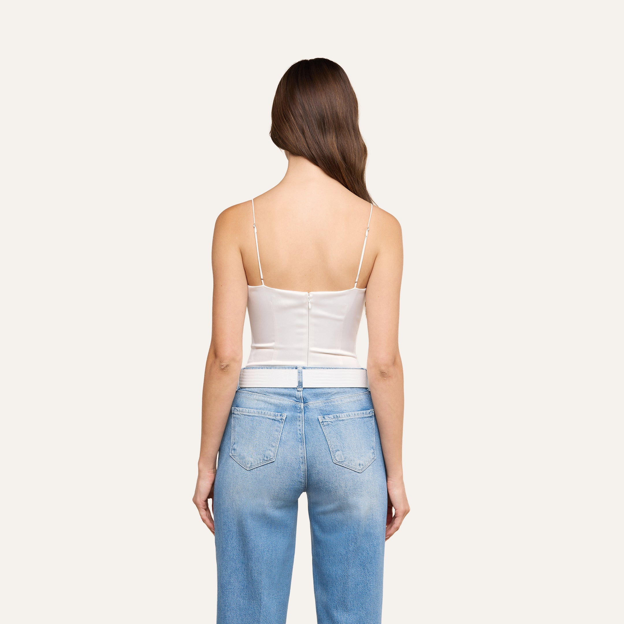 A woman with long brown hair stands facing away against a plain light background, wearing the Dara Stretch Satin Bodysuit in white with adjustable spaghetti straps and light blue jeans.
