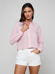 A woman stands against a plain light background, looking at the camera with one hand touching her collar, wearing the Cosette Shirt—a pink striped jacquard poplin button-up—paired with white shorts.