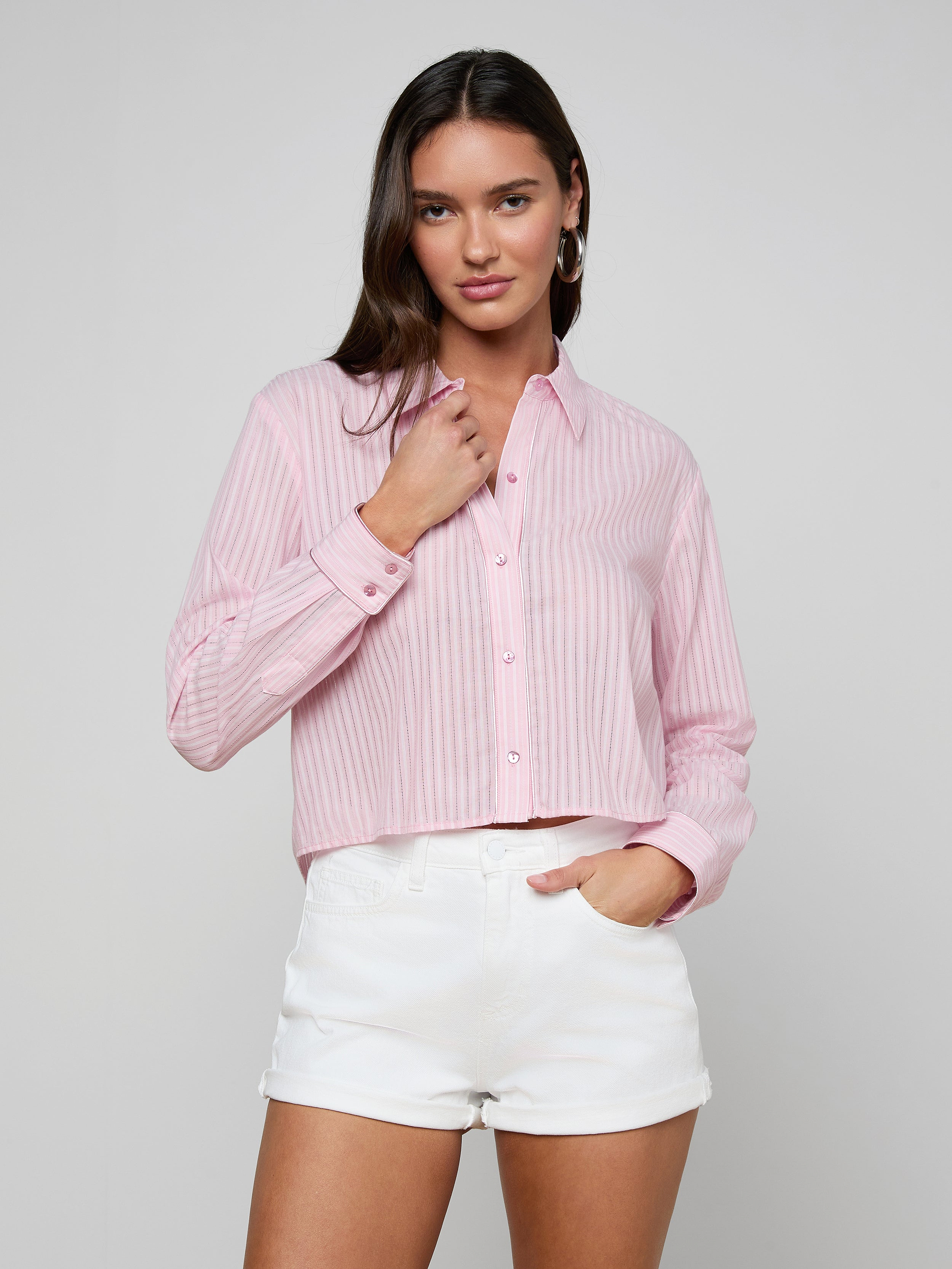 A woman stands against a plain light background, looking at the camera with one hand touching her collar, wearing the Cosette Shirt—a pink striped jacquard poplin button-up—paired with white shorts.