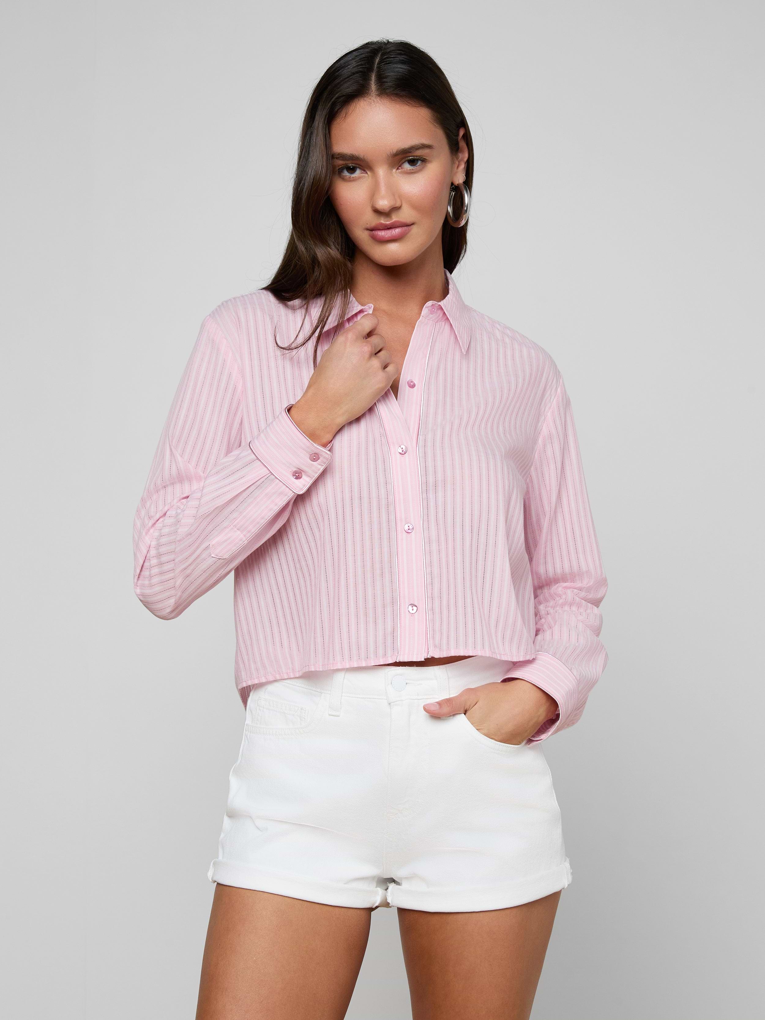 A woman stands against a plain light background, looking at the camera with one hand touching her collar, wearing the Cosette Shirt—a pink striped jacquard poplin button-up—paired with white shorts.