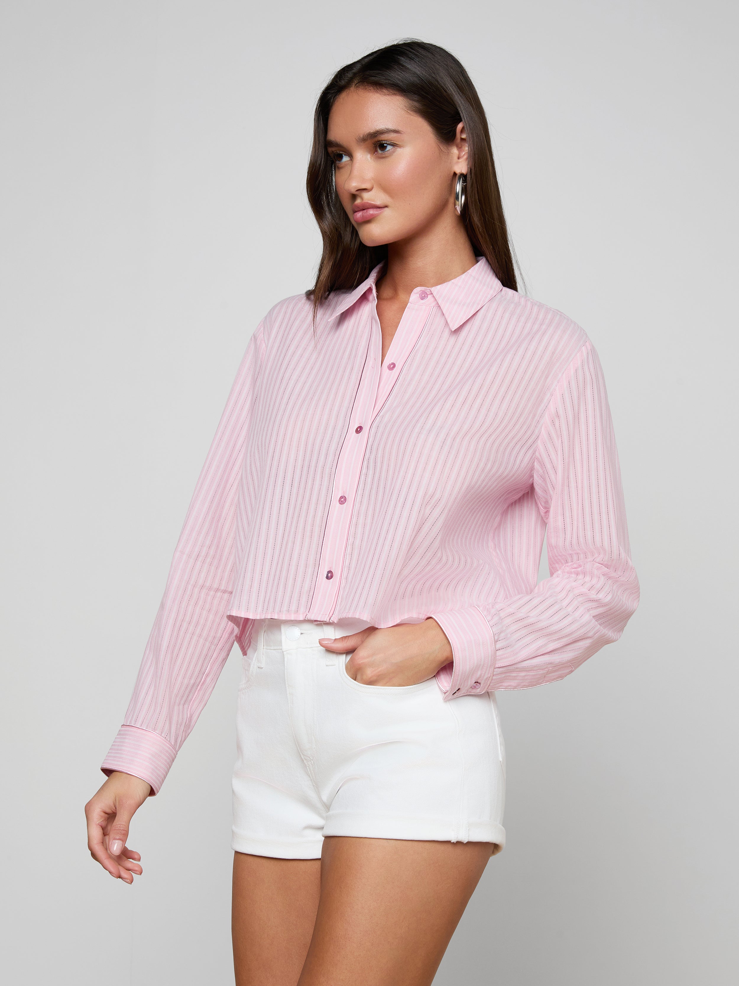 A woman wearing the Cosette Shirt—a light pink, long-sleeve shirt with white vertical stripes—paired with white high-waisted shorts, poses against a plain off-white background.