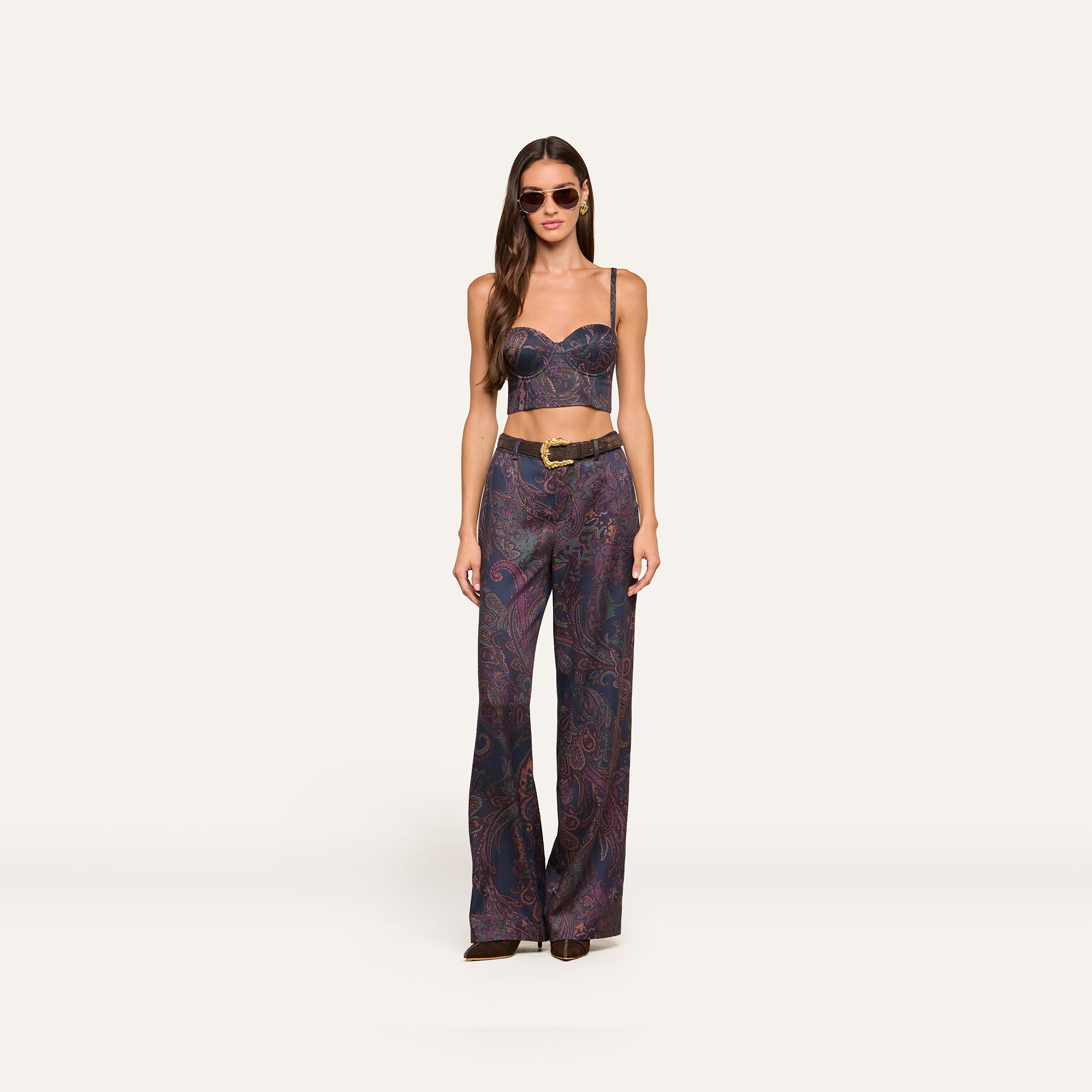 A woman wearing sunglasses and the Sonya Bustier—a navy paisley top with built-in support—paired with matching high-waisted, wide-leg pants featuring a belt. She stands facing forward against a plain light background.