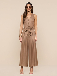 A woman wearing sunglasses and the sleeveless, tan Jianna Vest linen jumpsuit with a waist tie stands against a plain white background, her hands relaxed at her sides, accessorized with heeled sandals and bracelets.