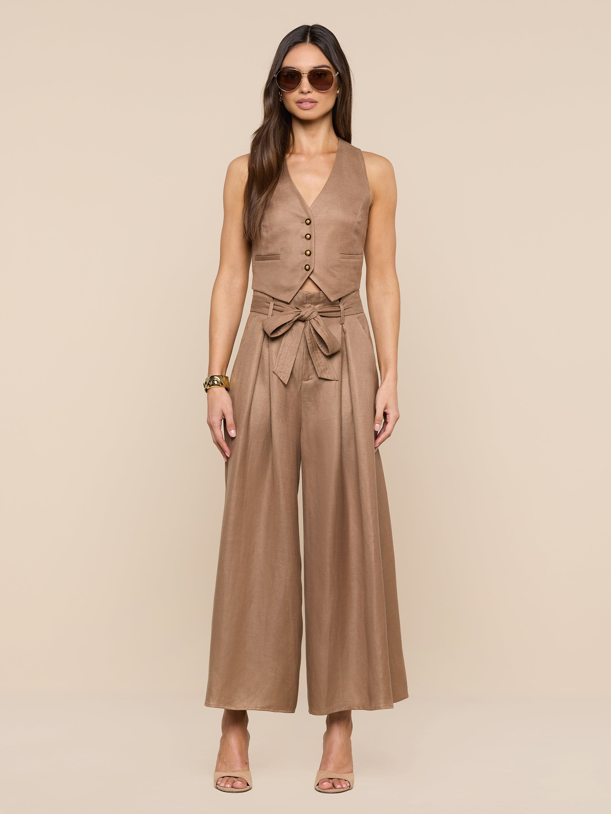 A woman wearing sunglasses and the sleeveless, tan Jianna Vest linen jumpsuit with a waist tie stands against a plain white background, her hands relaxed at her sides, accessorized with heeled sandals and bracelets.
