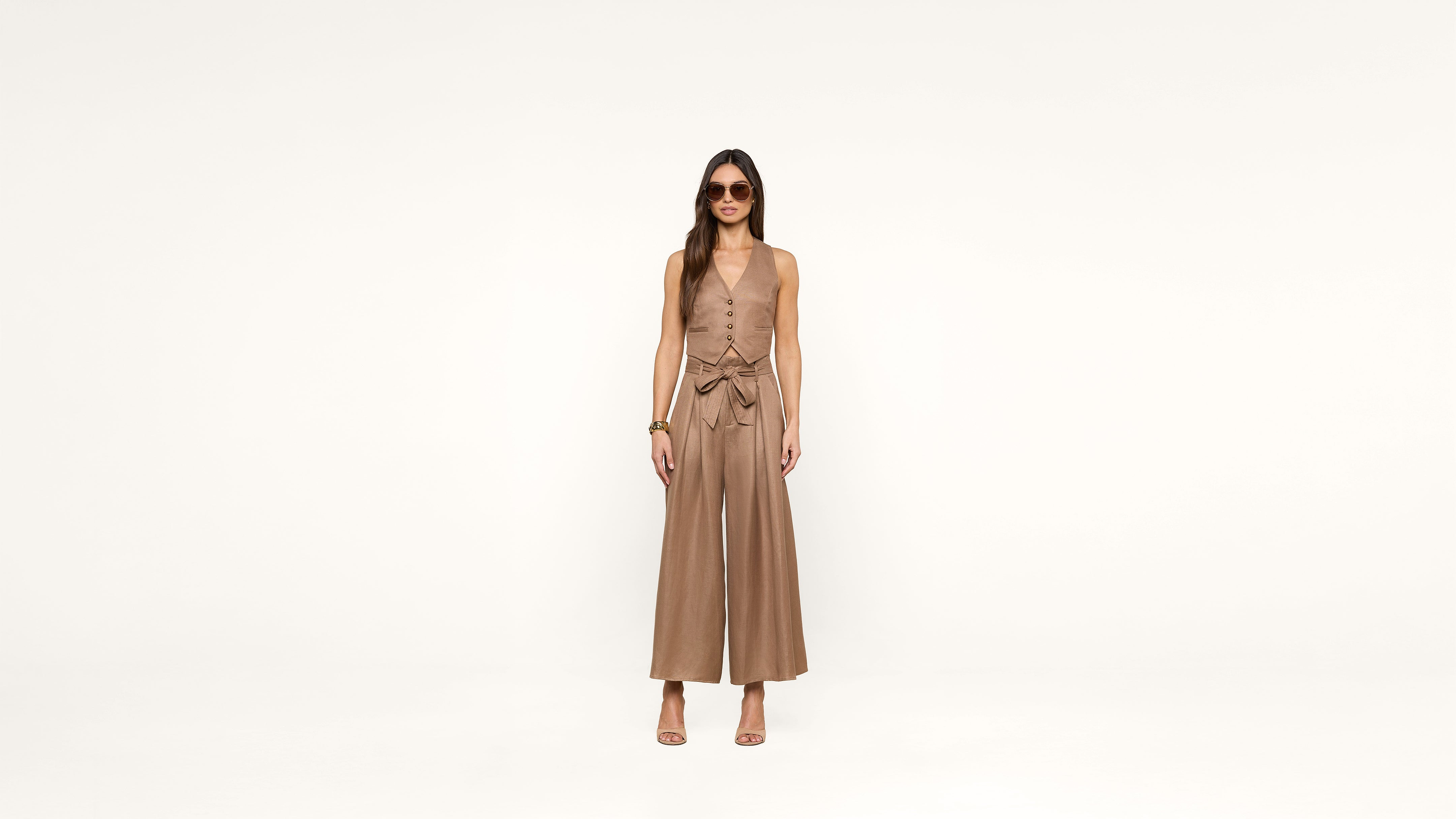 Woman stands against a plain white background, wearing a tan sleeveless buttoned jumpsuit with wide legs, tan high-heeled sandals, sunglasses, and a bracelet.