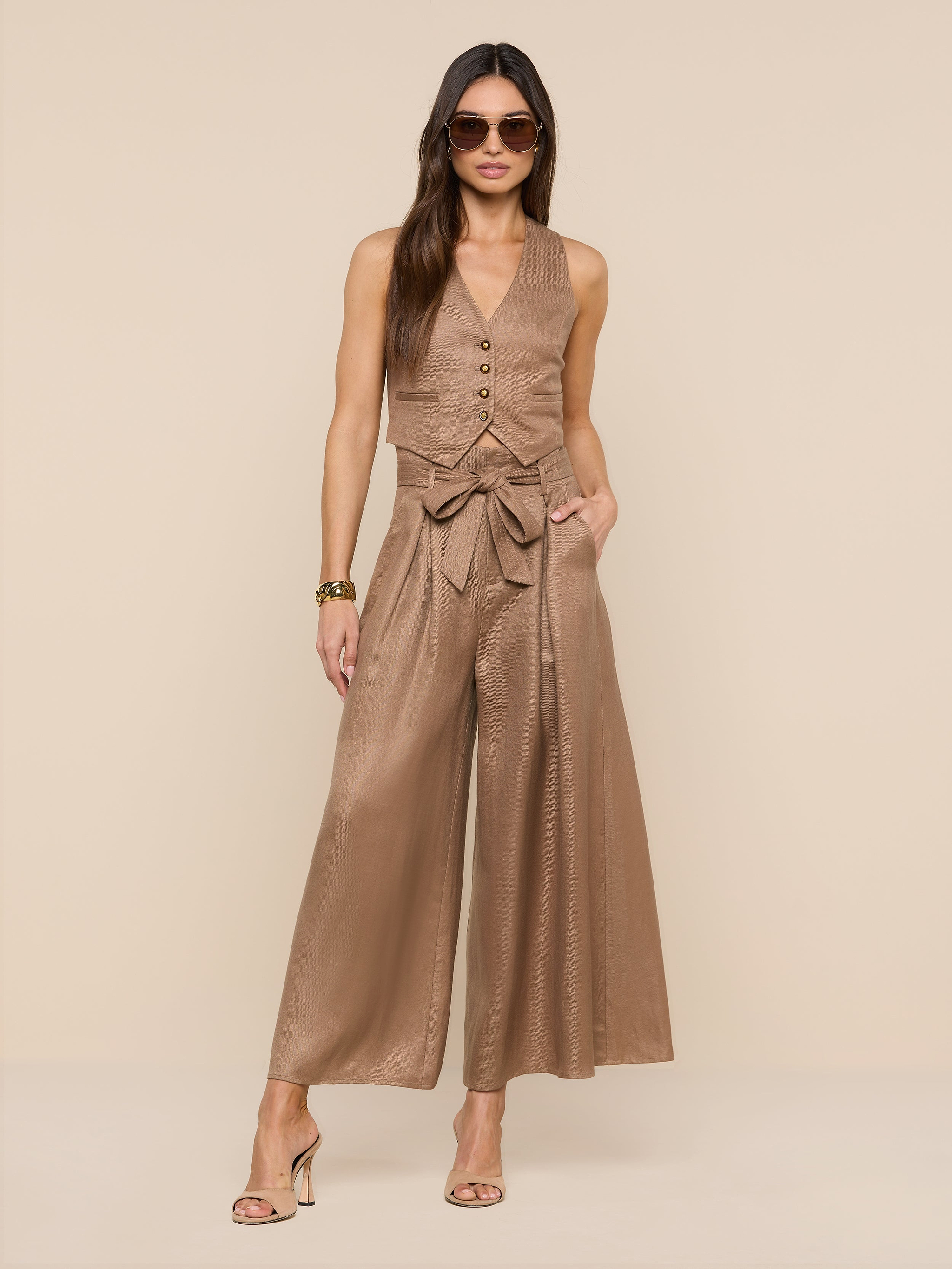 Woman wearing sunglasses, the Jianna Vest—a sleeveless linen style—wide-leg high-waisted pants with a bow belt, and heeled sandals stands against a plain white background.