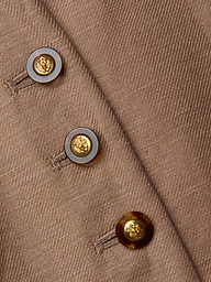 Close-up of the Jianna Vest—a brown cropped vest featuring three round buttons. Each button has a gold center with a raised emblem, encircled by a dark brown ring, and they are sewn in a diagonal line.