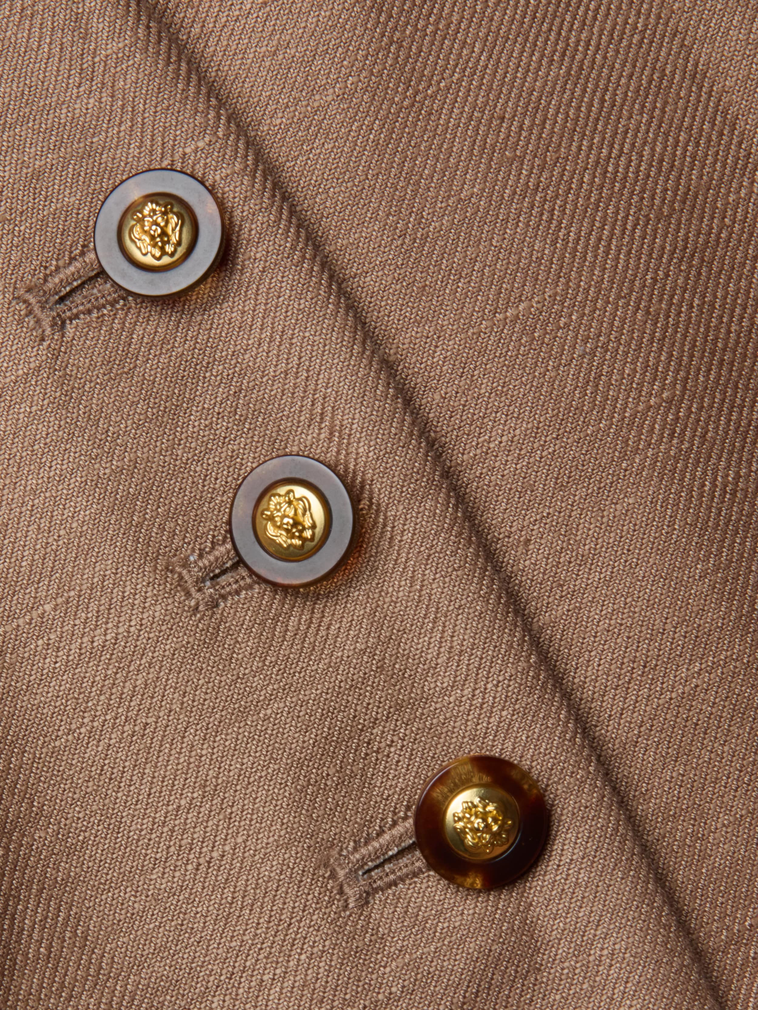 Close-up of the Jianna Vest—a brown cropped vest featuring three round buttons. Each button has a gold center with a raised emblem, encircled by a dark brown ring, and they are sewn in a diagonal line.